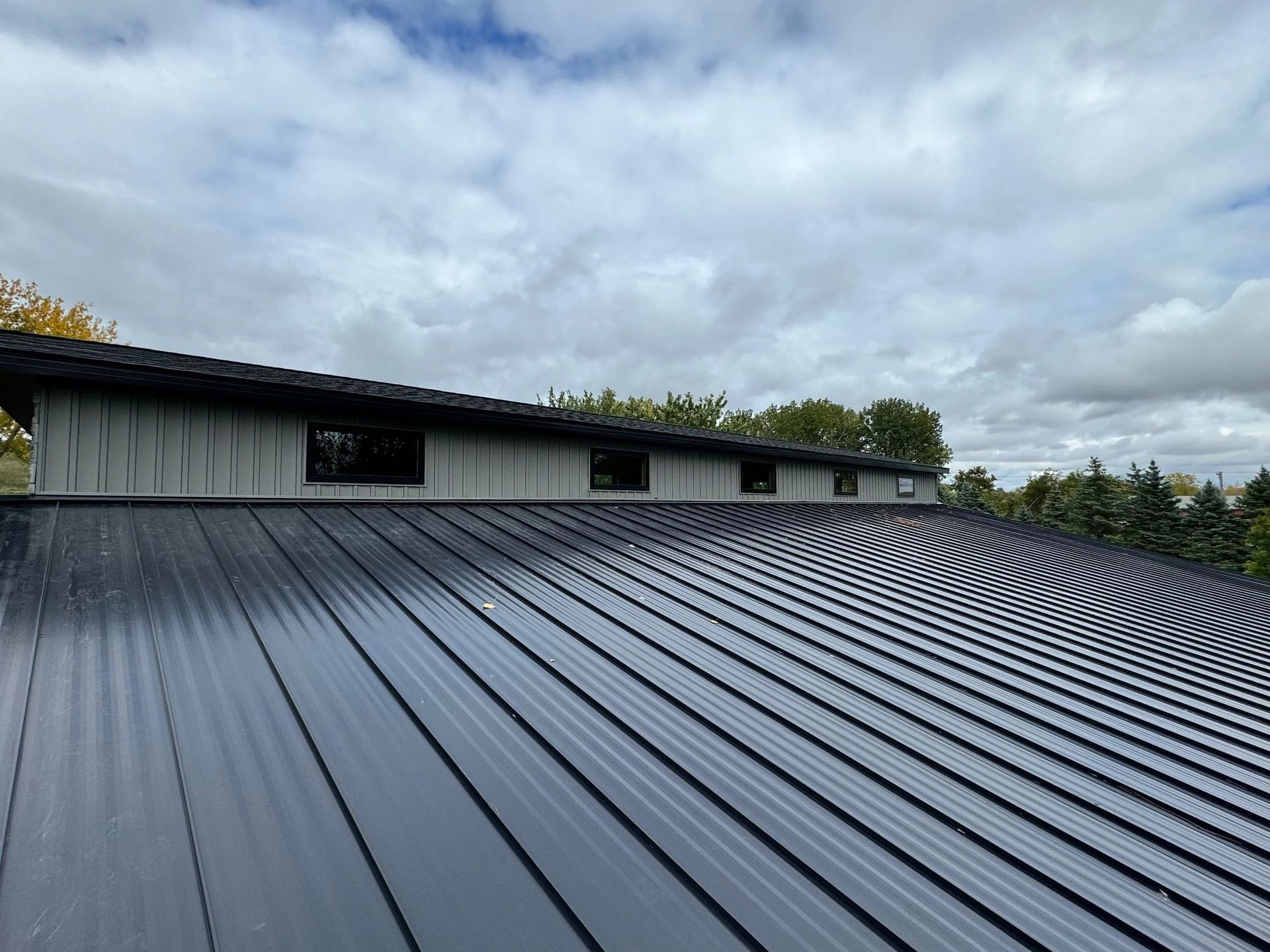 Dark gray metal roof with long ridges beneath a cloudy sky and tree line