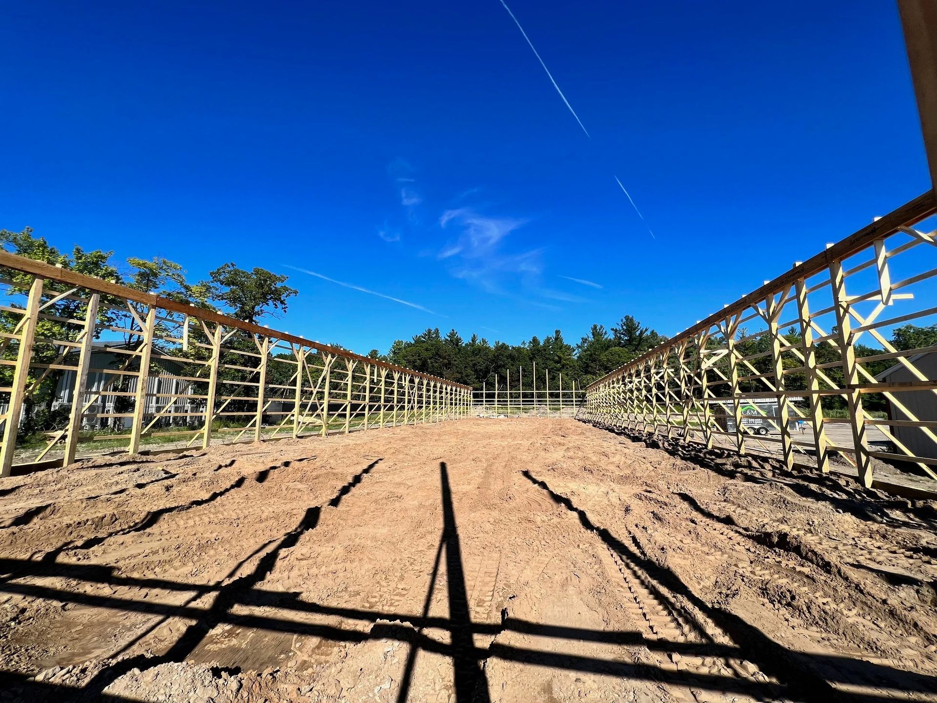 Dirt path between wooden fence rails under a bright blue sky, with trees in the distance.