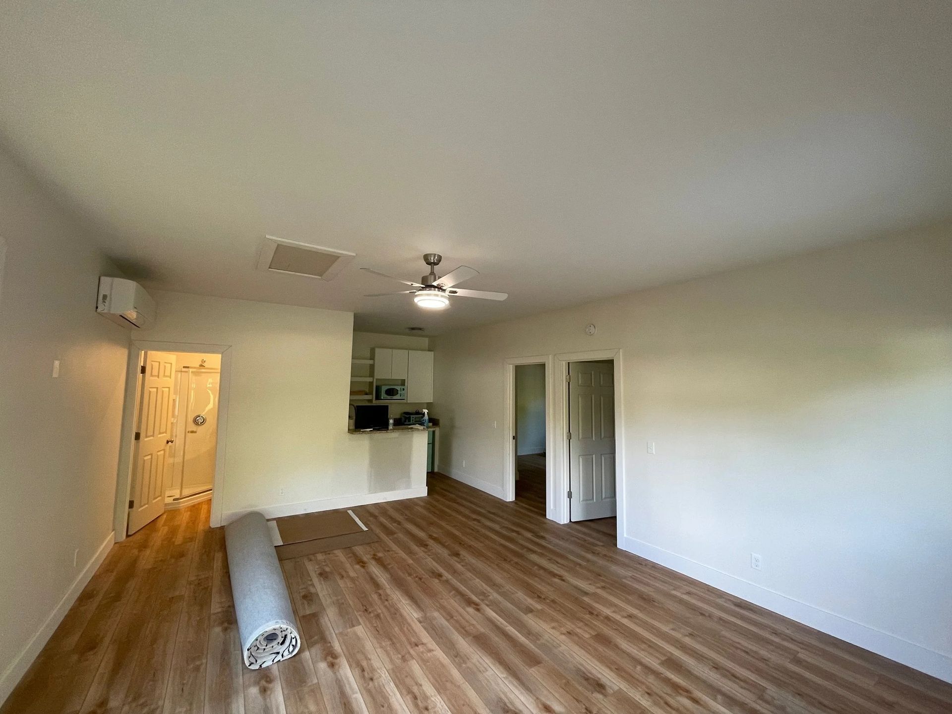 Empty living room with wood floors, white walls, ceiling fan, and rolled carpet on the floor