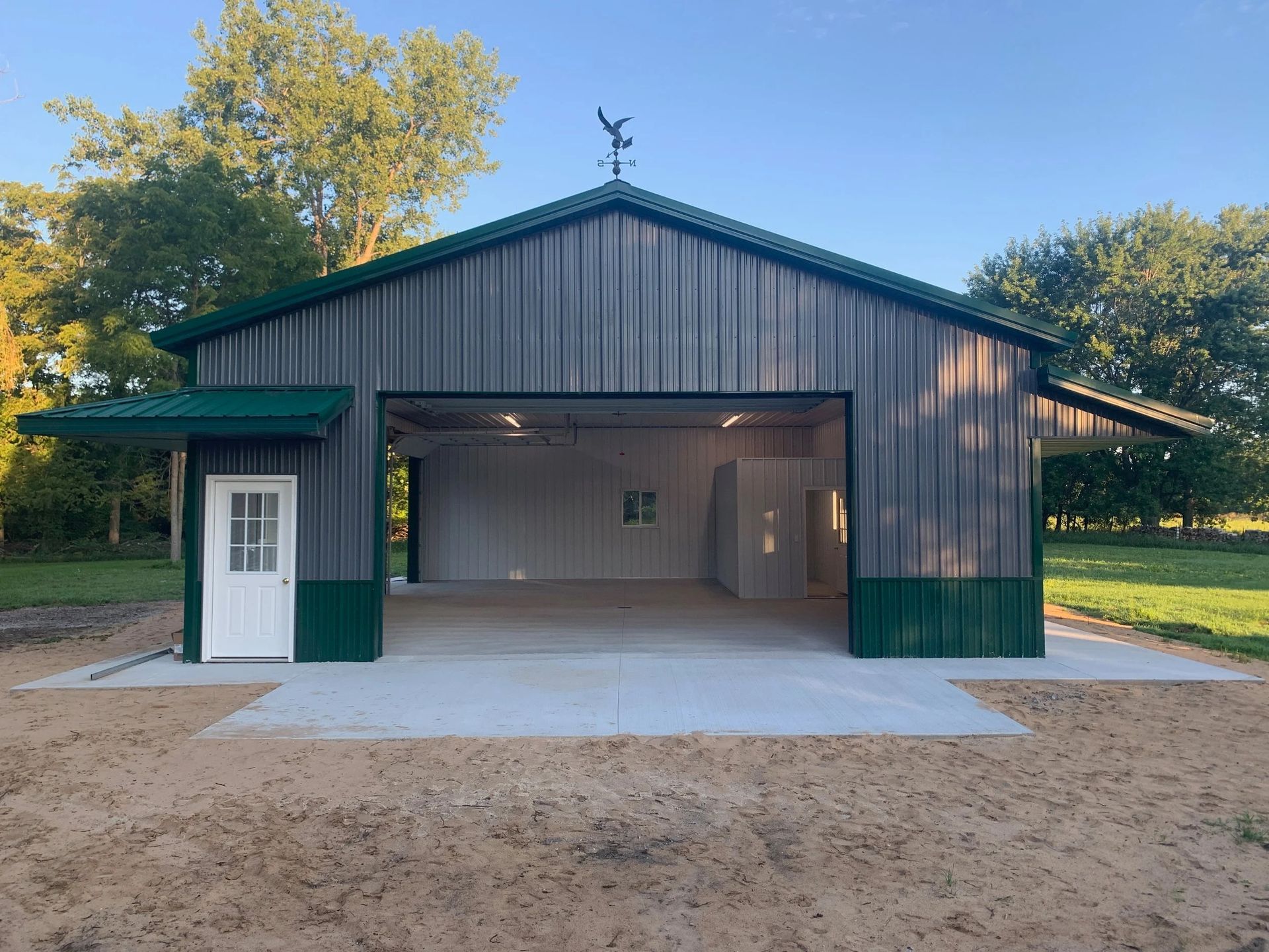 Green metal barn with open garage bay and white side door on a dirt lot.