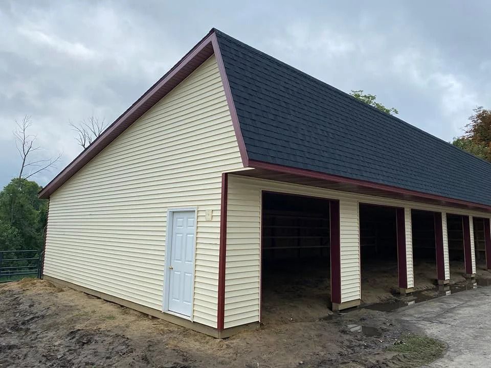 Cream-sided barn with a dark roof and open bays on a dirt lot under cloudy skies