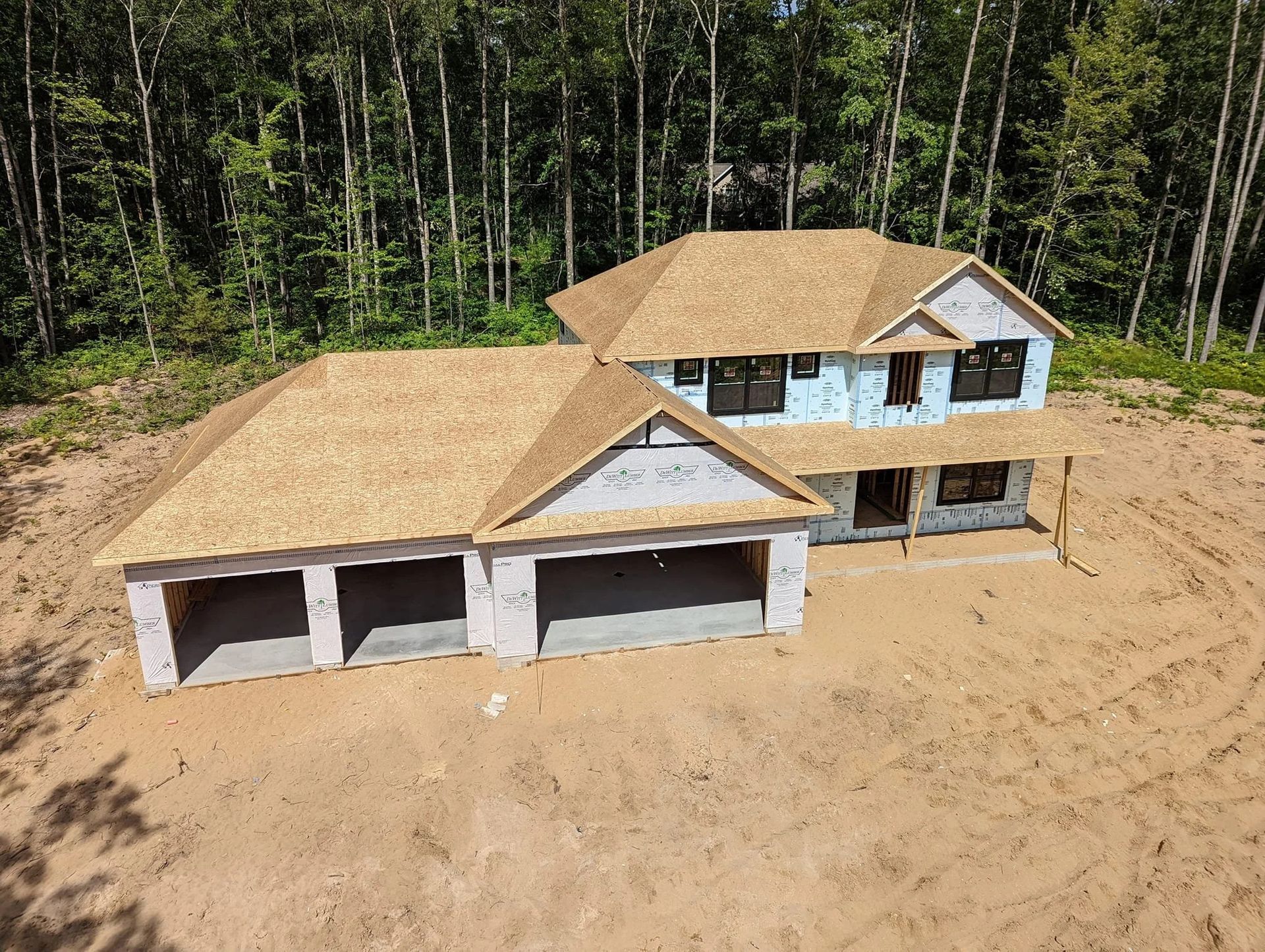 New house under construction on a sandy lot beside a wooded area, with attached garage and unfinished exterior