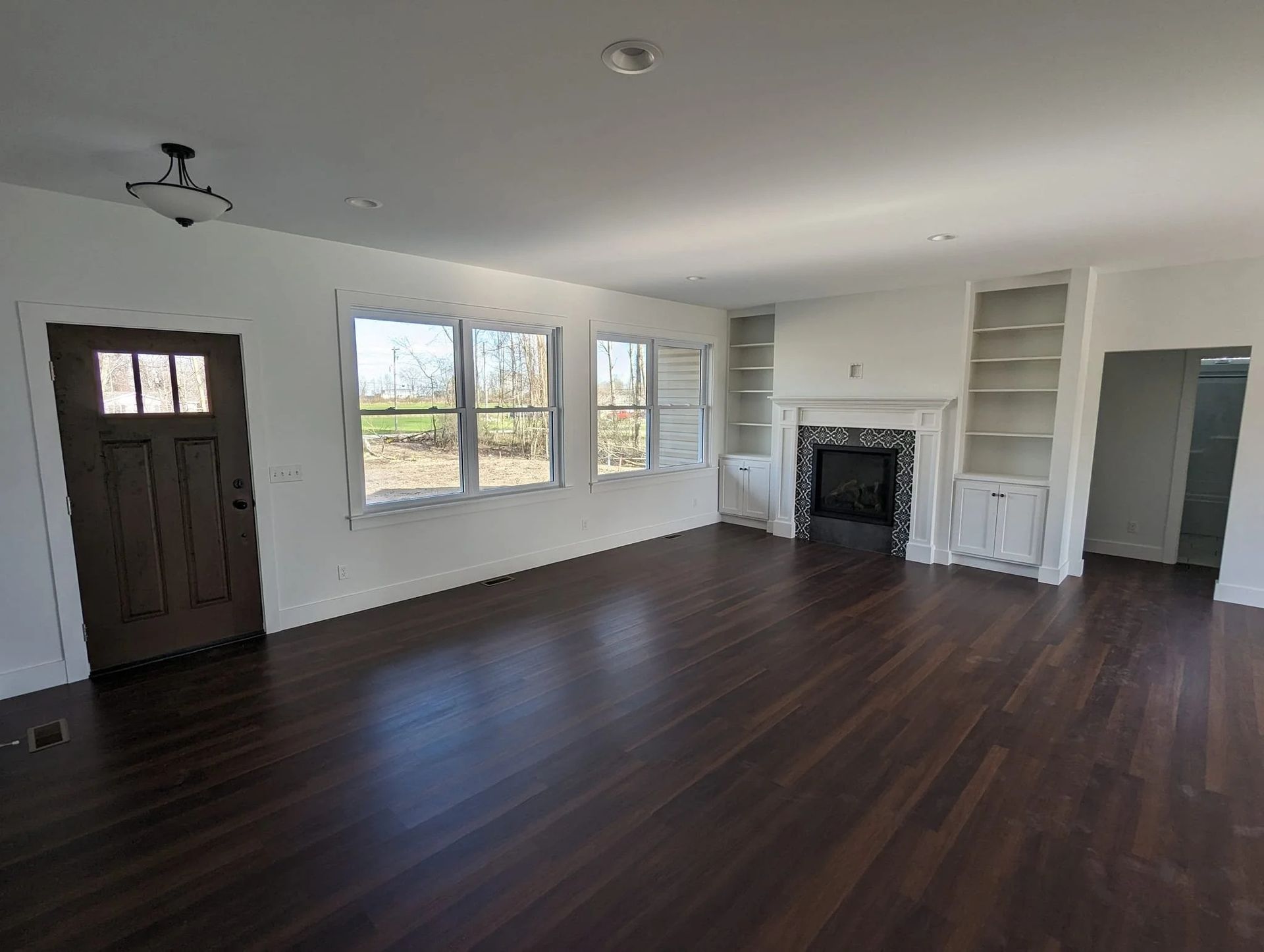 Empty living room with dark wood floors, white walls, large windows, and a fireplace.
