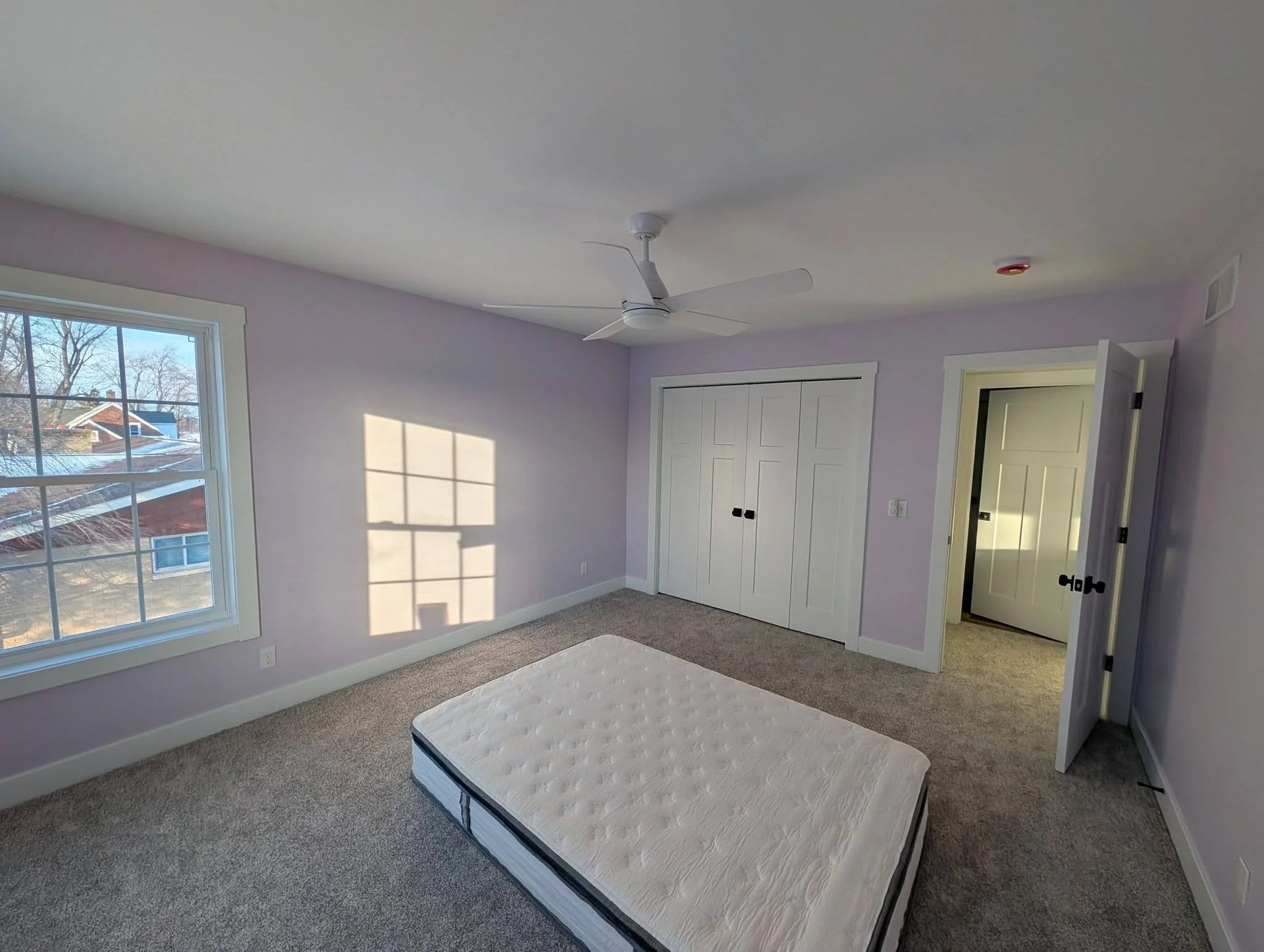 Empty lavender bedroom with a bed, closet doors, ceiling fan, and sunlight through a window.