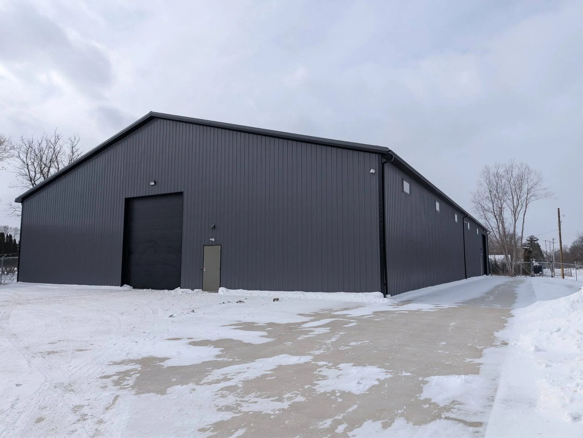 Dark gray industrial warehouse on a snowy lot under a cloudy sky