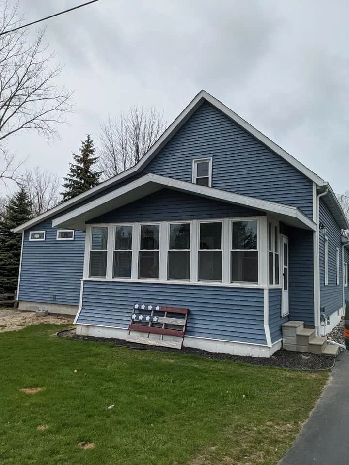 Blue house with white trim and enclosed porch beside a driveway under cloudy skies