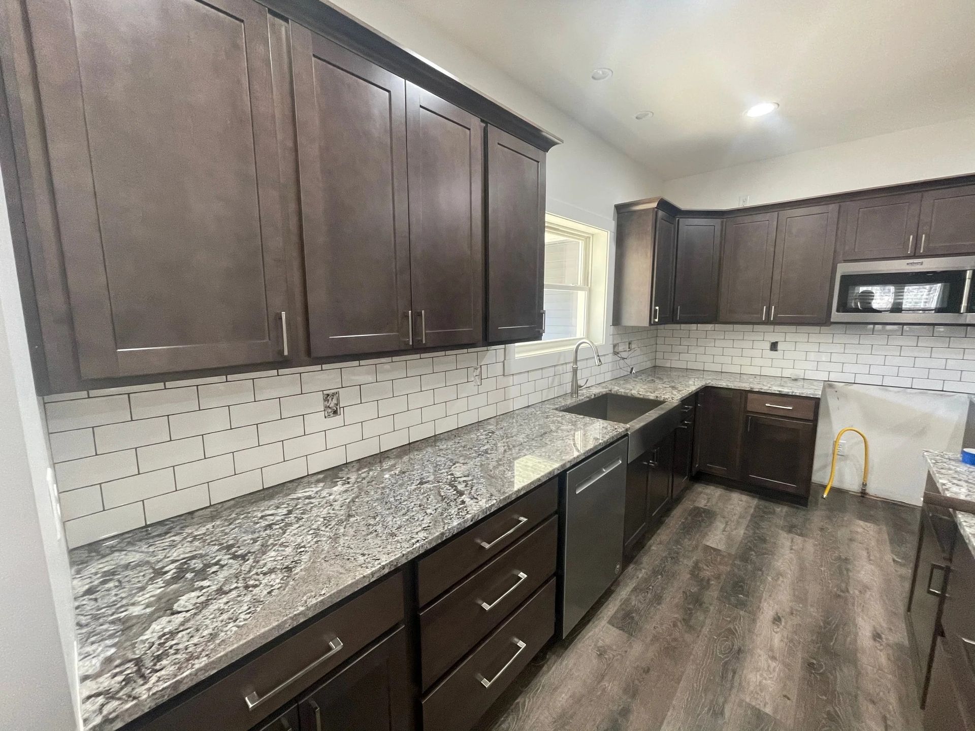 Modern kitchen with dark wood cabinets, granite counters, white tile backsplash, and wood-look flooring.