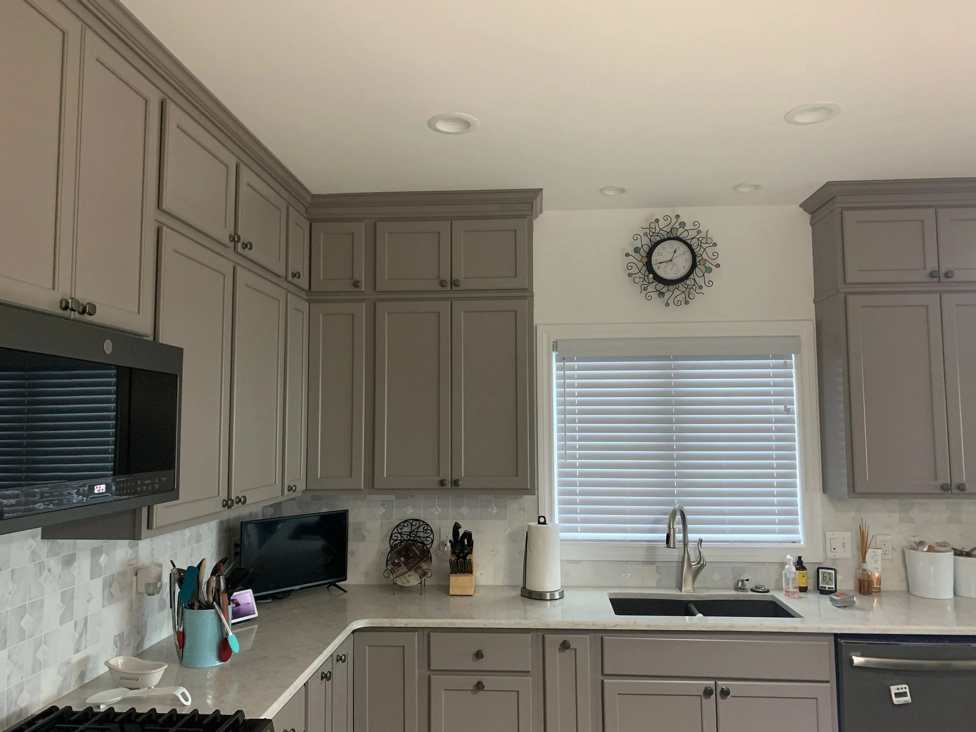Modern kitchen with gray cabinets, white counters, sink, and a window with blinds and wall clock.