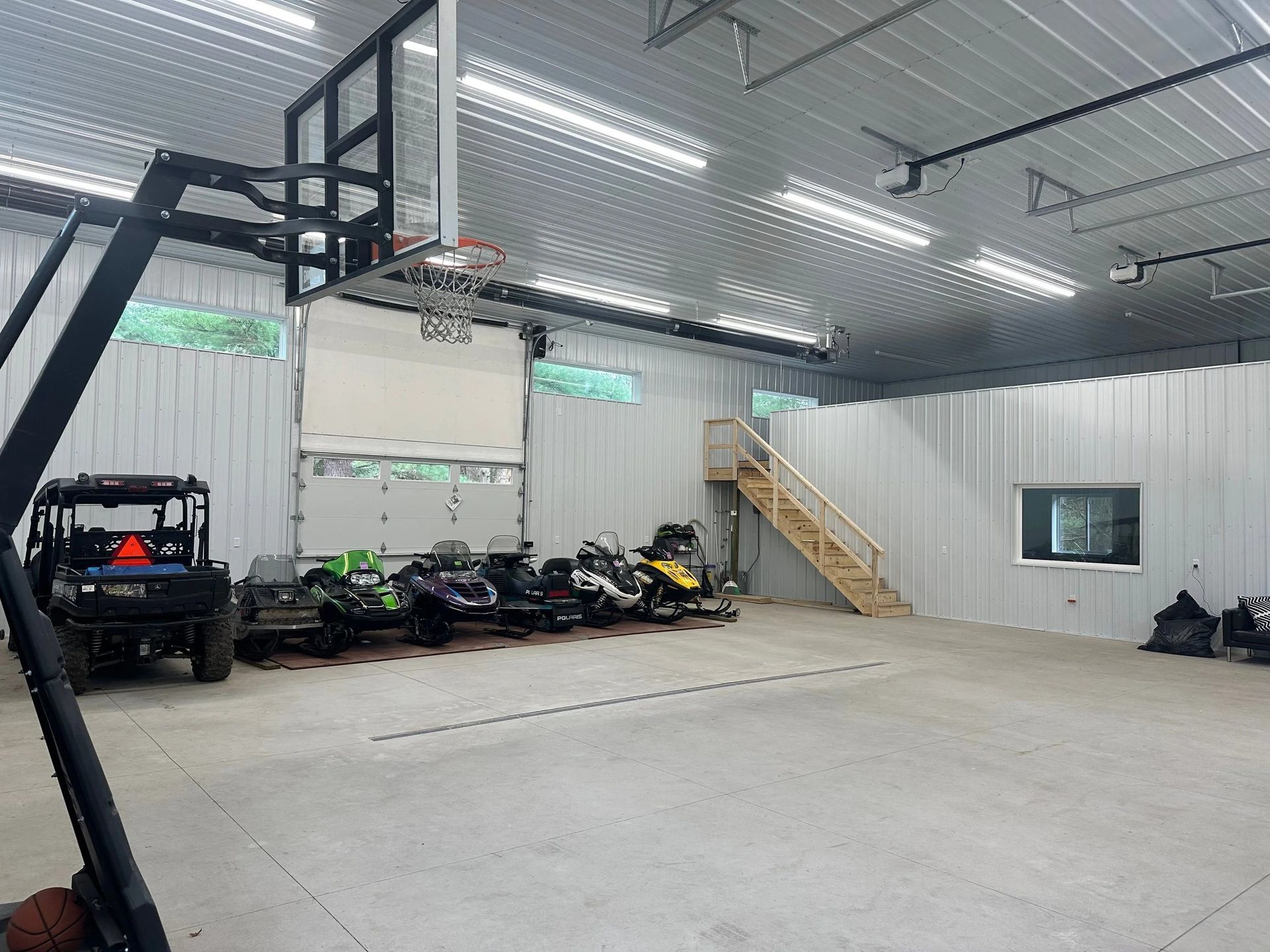 Indoor garage with concrete floor, basketball hoop, motorcycles, and a wooden staircase against white walls