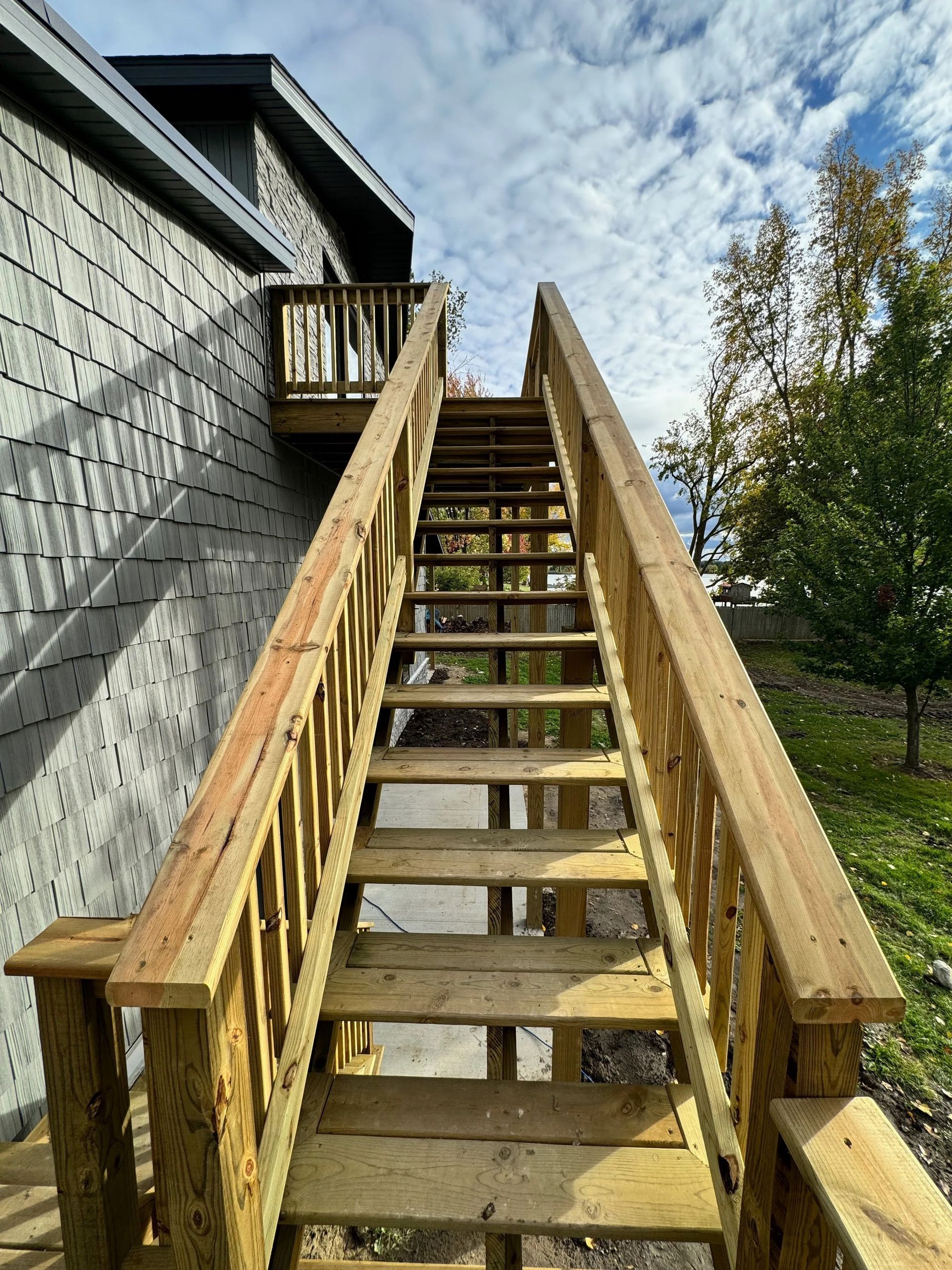 Wooden outdoor staircase beside a gray house, leading up to a deck under a cloudy sky.