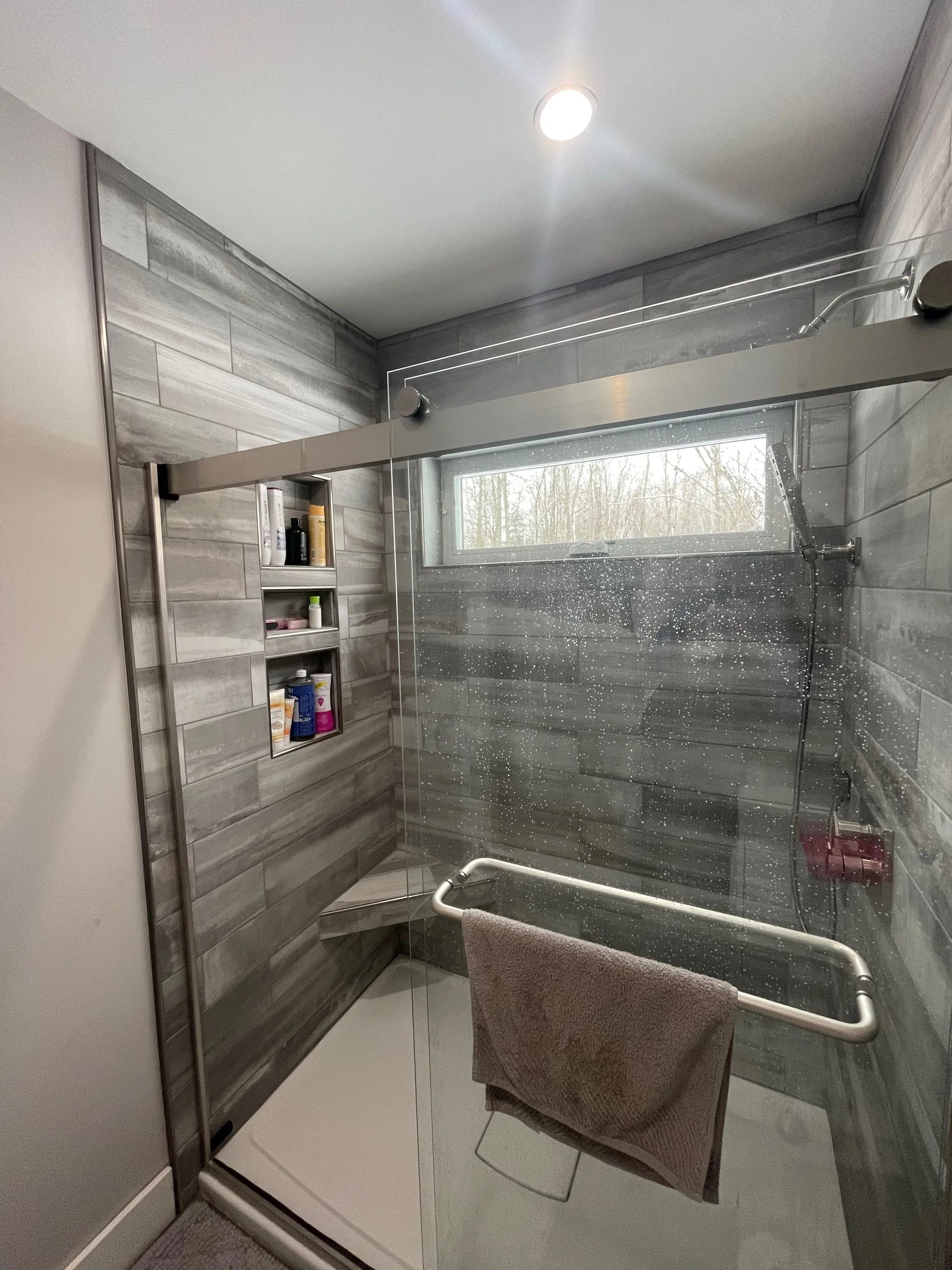 Modern shower with glass enclosure, built-in shelves, towel bar, and gray tiled walls under a bright light