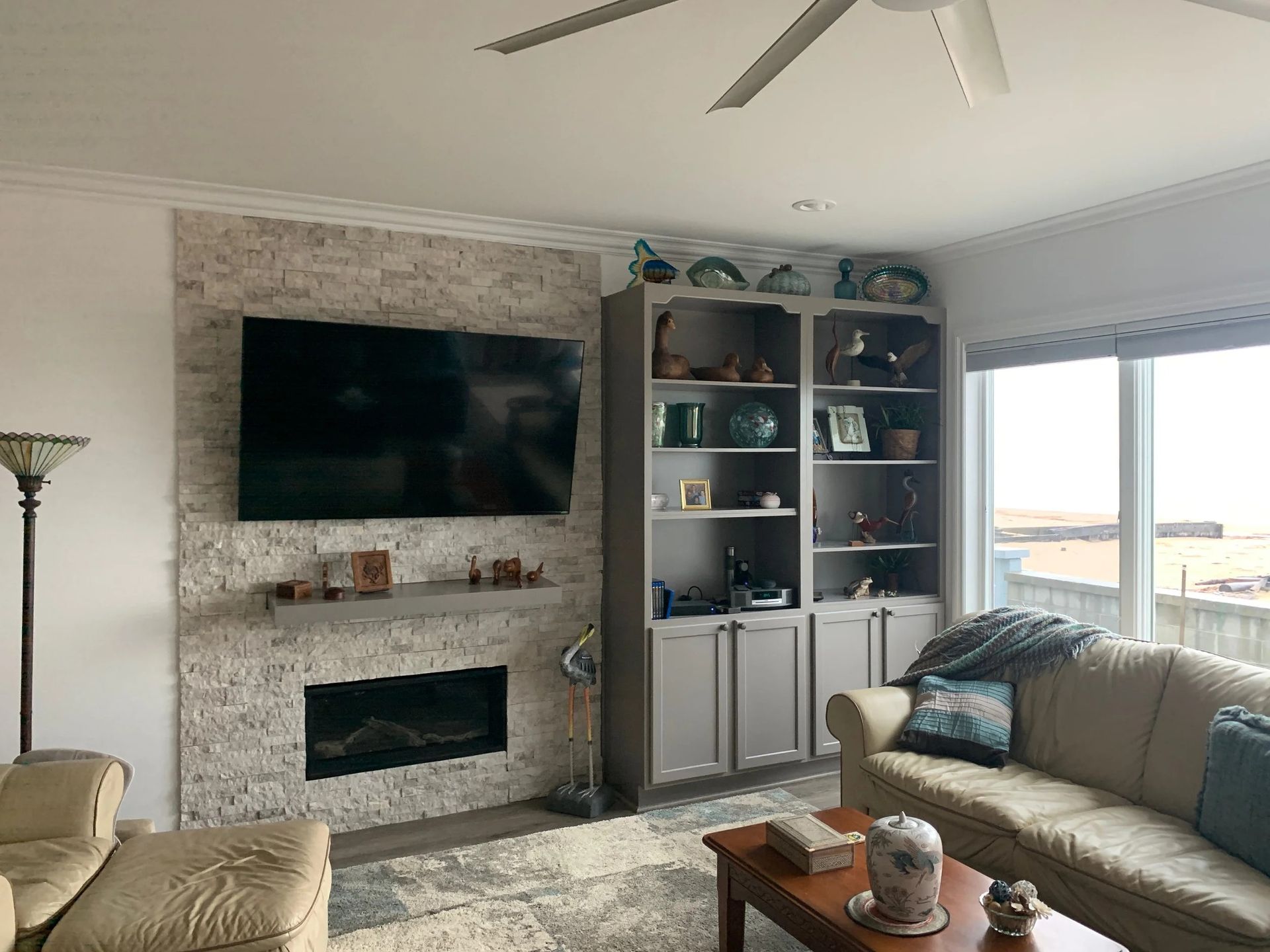 Modern living room with wall-mounted TV, fireplace, built-in shelves, and a beige couch by a large window