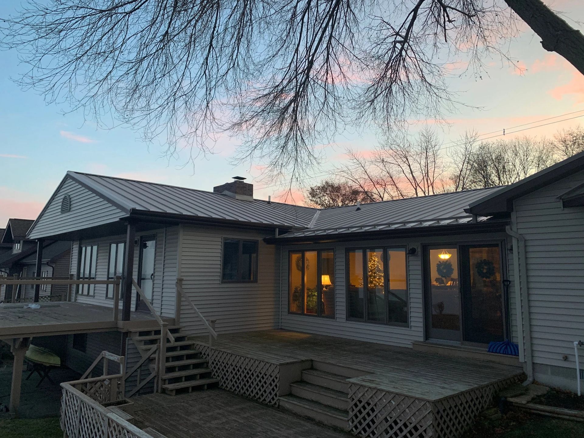 Wooden house with a porch and lit windows at dusk, framed by bare tree branches.