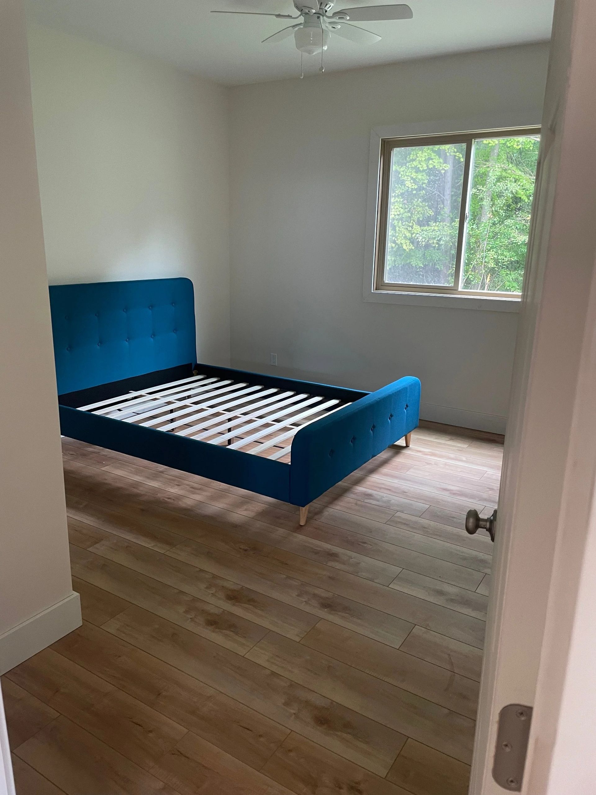 Empty bedroom with a blue bed frame, hardwood floor, and a window overlooking trees.