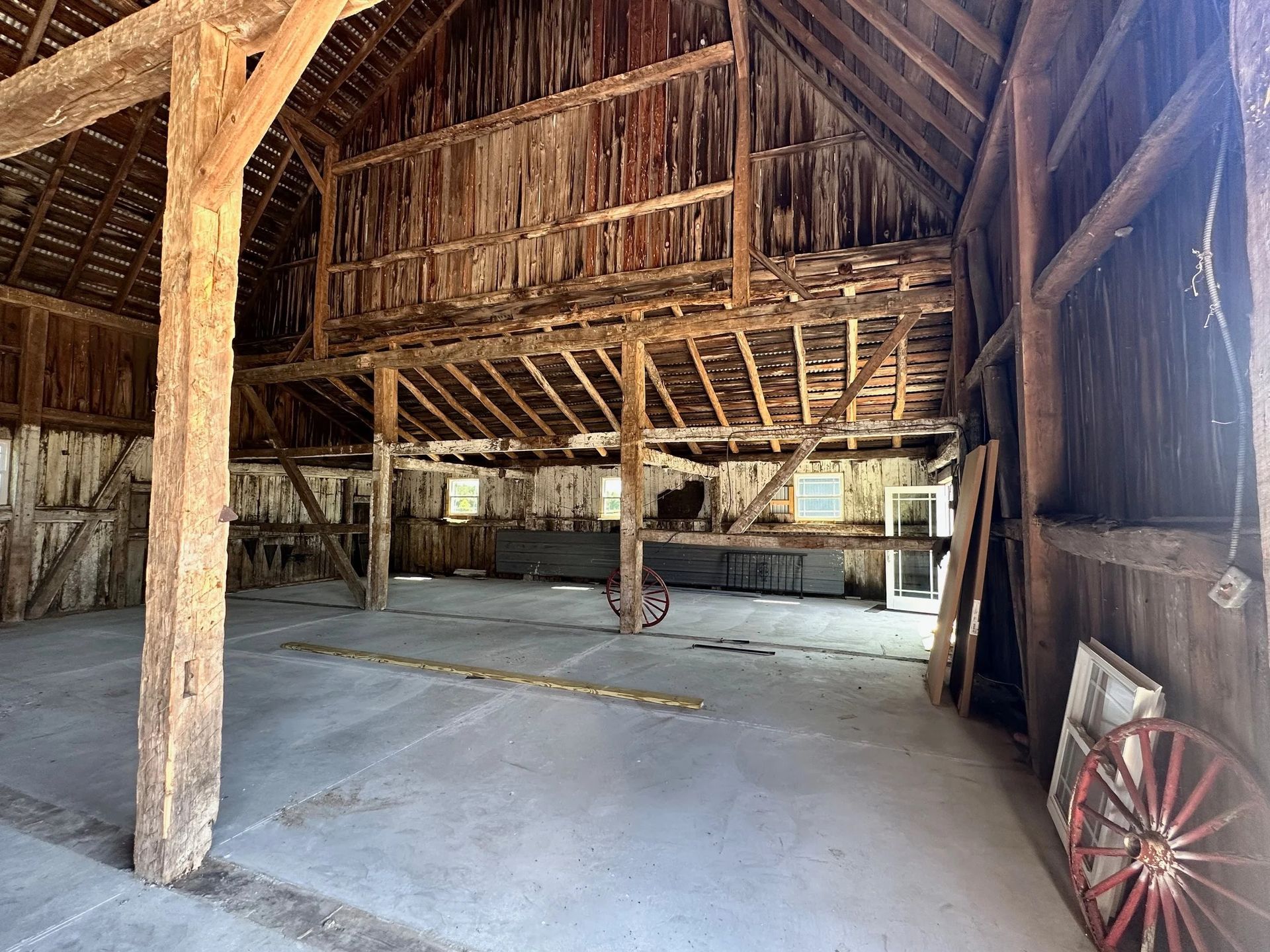 Inside a rustic wooden barn with a concrete floor, hayloft, and a red wagon wheel on the right