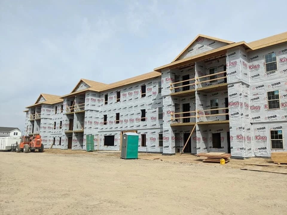 Row of unfinished townhouses under construction, wrapped in white sheathing on a sandy lot.