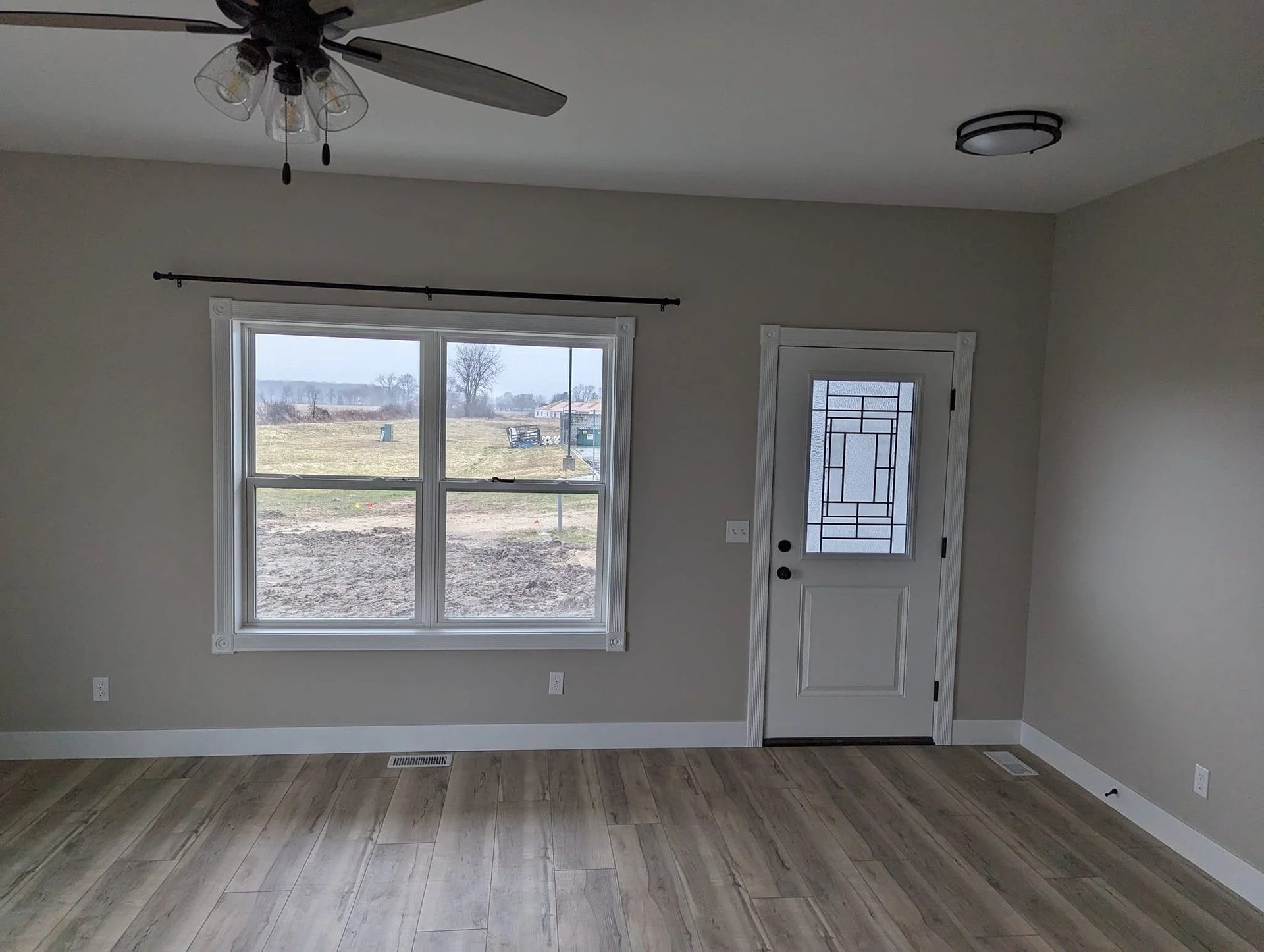 Empty living room with gray walls, wood-look floor, ceiling fan, large window, and front door.