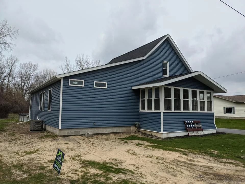 Blue house with white-trimmed windows and porch on a cloudy day, with a small sign in the yard