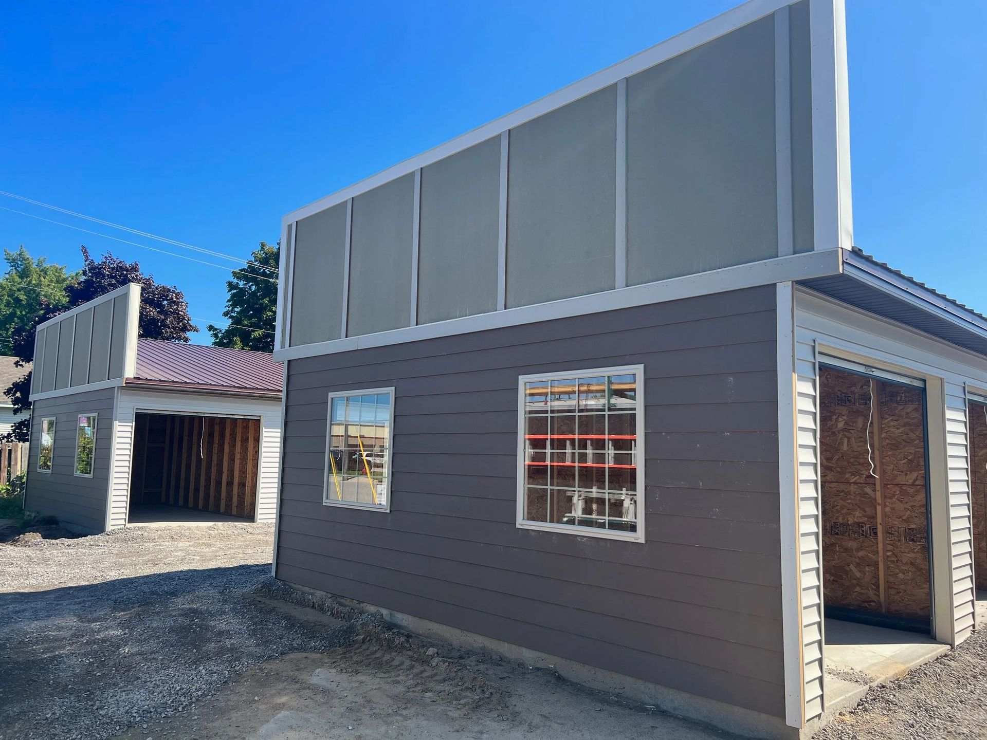 Modern gray building with large upper panels, windows, and a side opening on a gravel lot under a blue sky