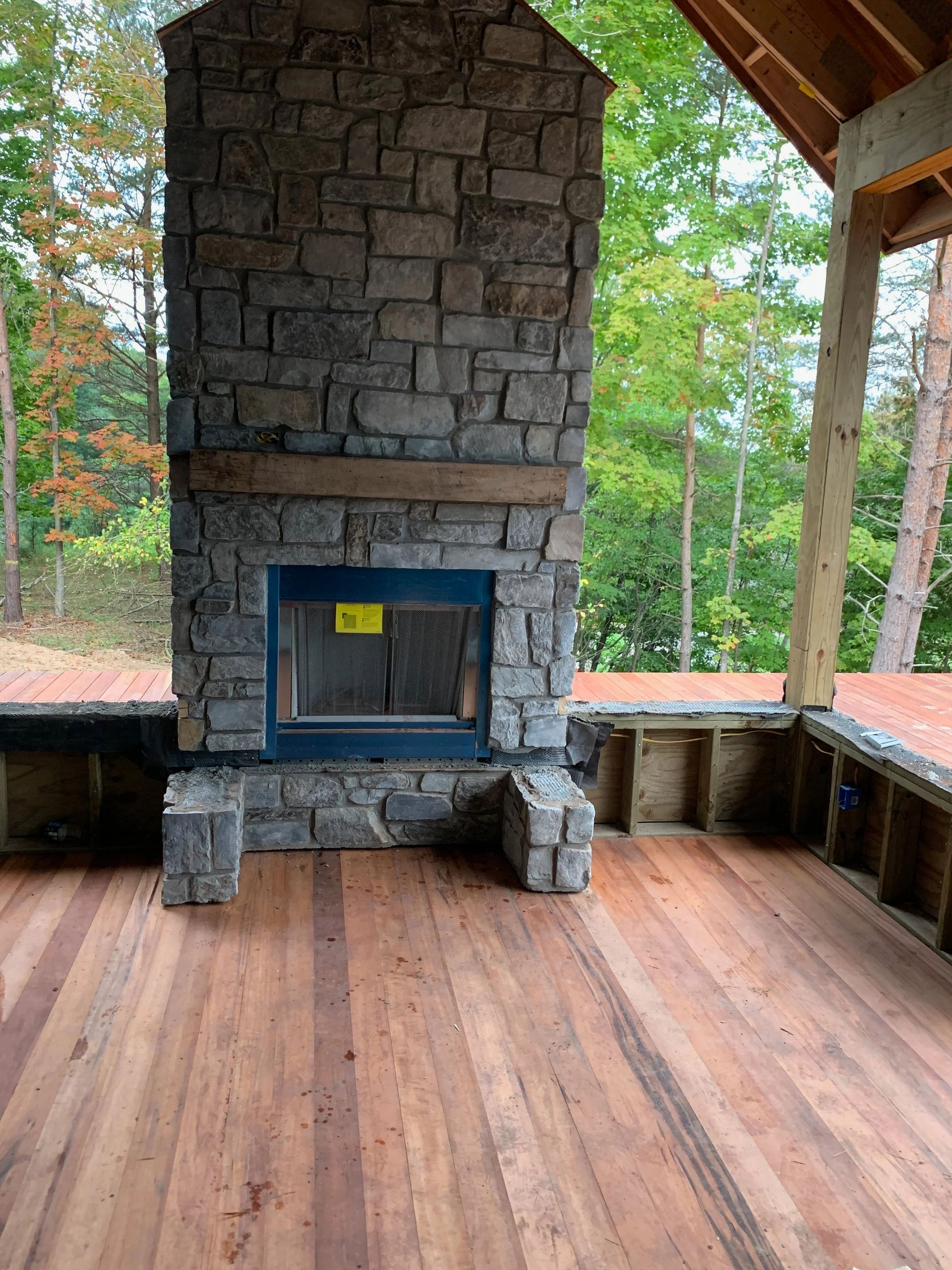 Stone outdoor fireplace on a wooden porch with a roof and trees in the background
