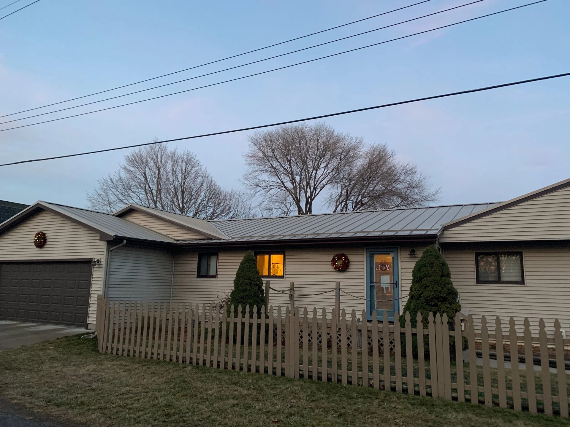 Single-story house with lit window, picket fence, garage, and leafless trees under a dusk sky