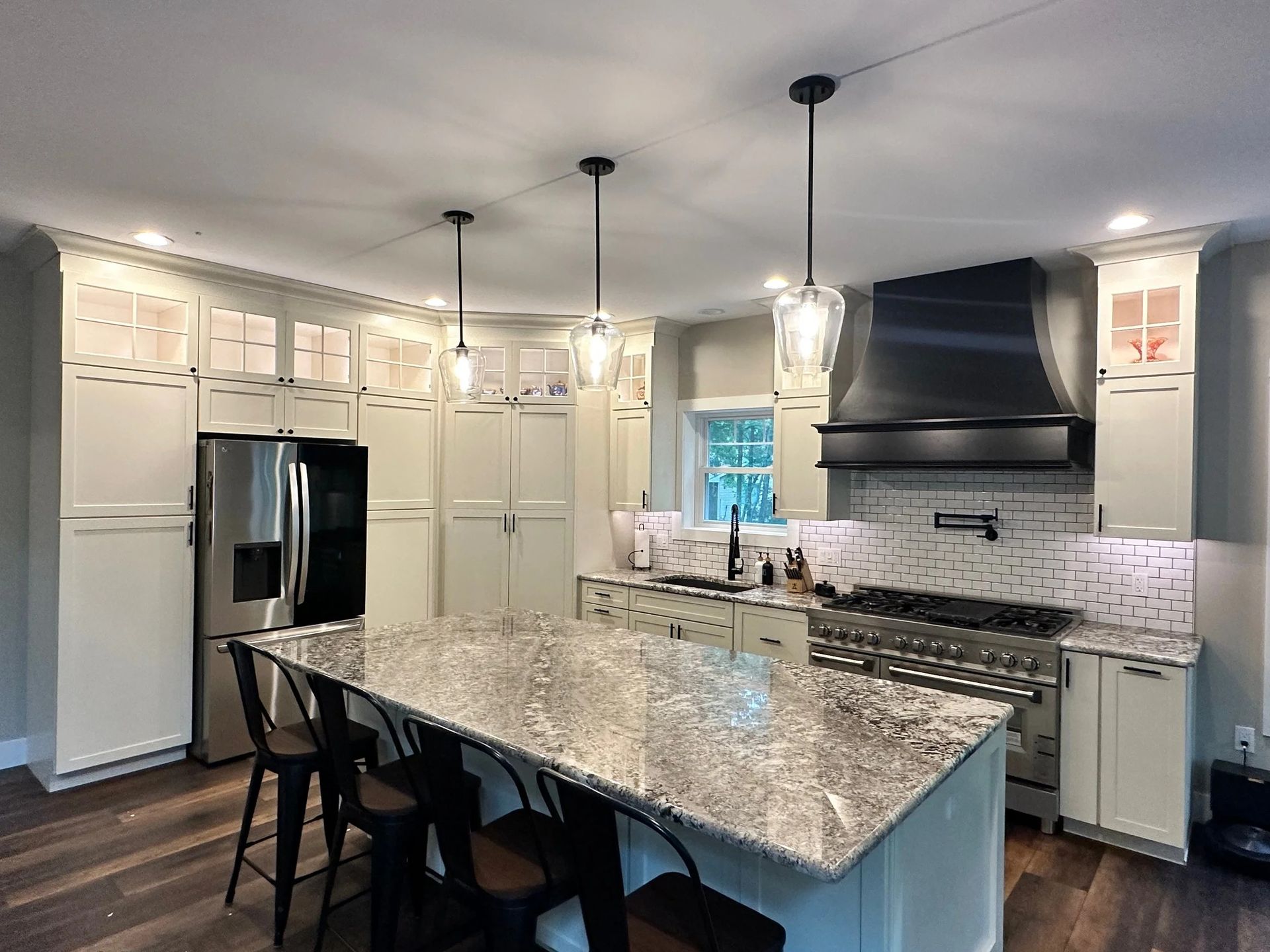 Modern white kitchen with granite island, pendant lights, black range hood, and stainless steel appliances