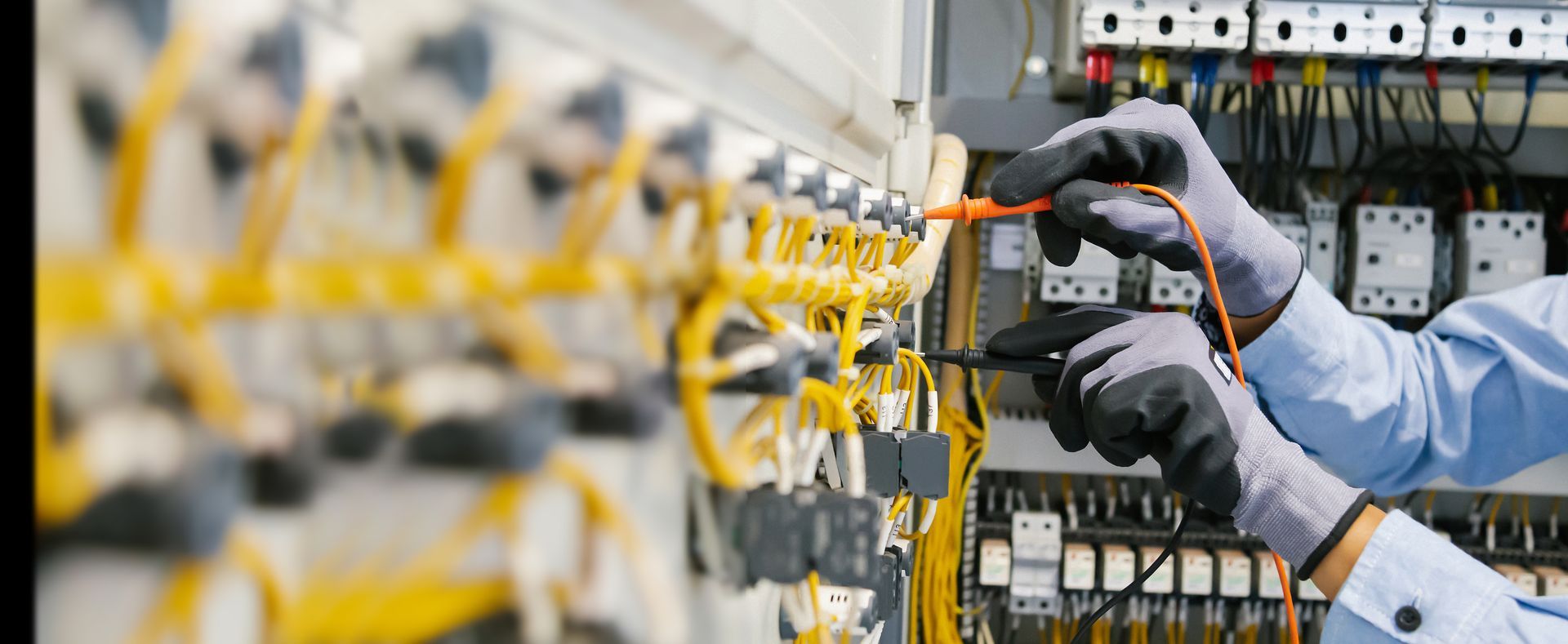 Un technicien portant des gants utilise des sondes de multimètre pour tester les connexions d'un tableau électrique rempli de fils jaunes.
