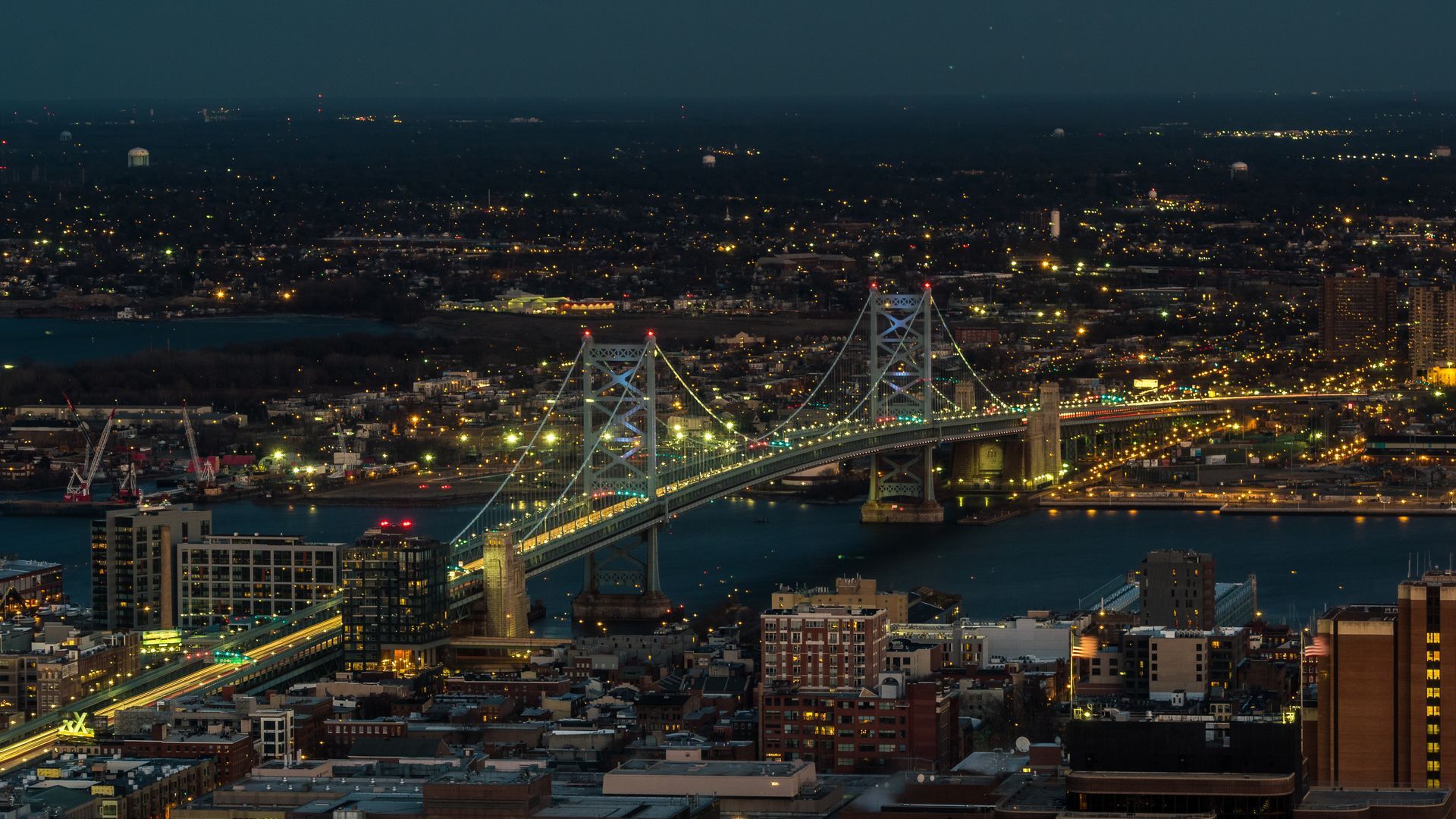 Nighttime cityscape with a lit suspension bridge spanning a river under city lights.