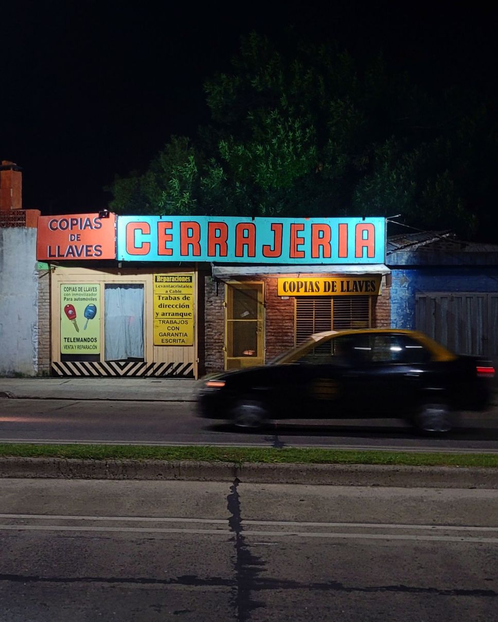 Fachada de tienda con un gran letrero de “CERRAJERIA” sobre un coche plateado aparcado.