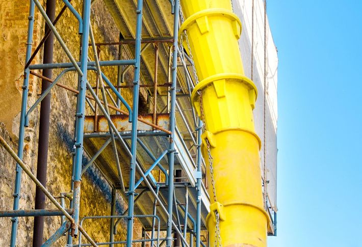 Yellow industrial chute beside blue scaffolding and a wall, with sky in the background