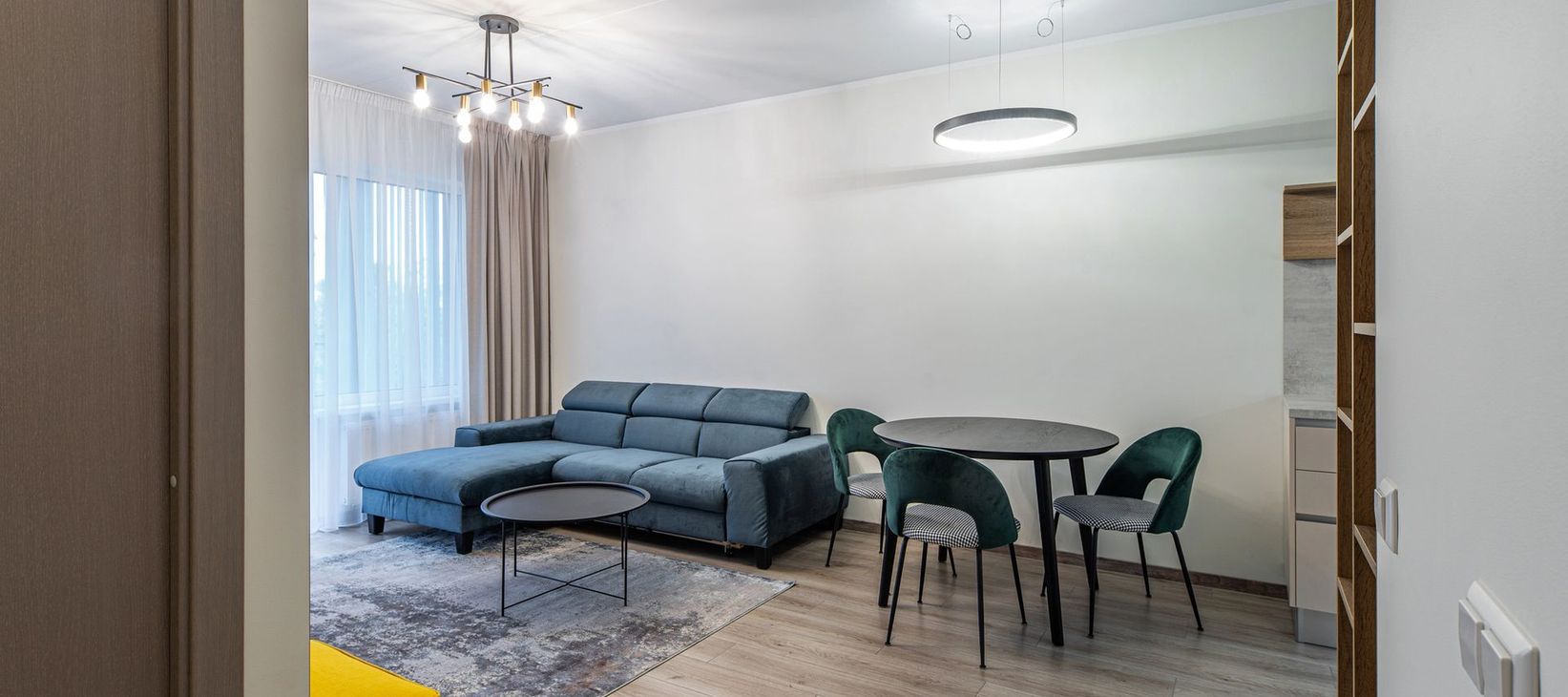 Bright living room with a blue sectional sofa, dining table, chairs, chandelier, and large window curtains.