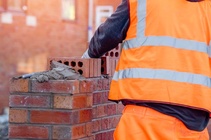 Construction worker laying red bricks on a wall, wearing an orange safety vest and gloves.