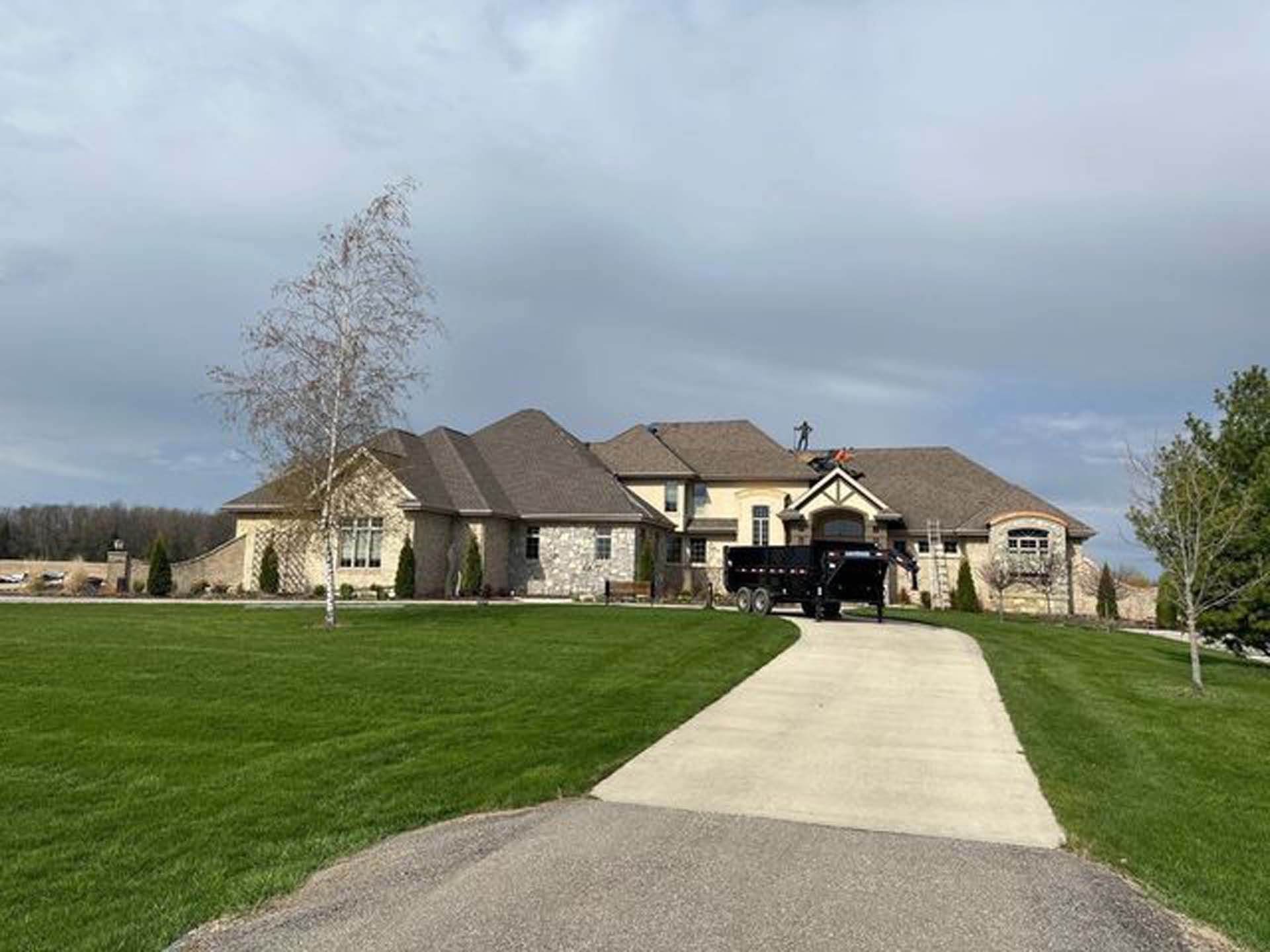 A large stone house with a multi-gabled brown roof, situated at the end of a concrete driveway on a sunny, grassy lawn.