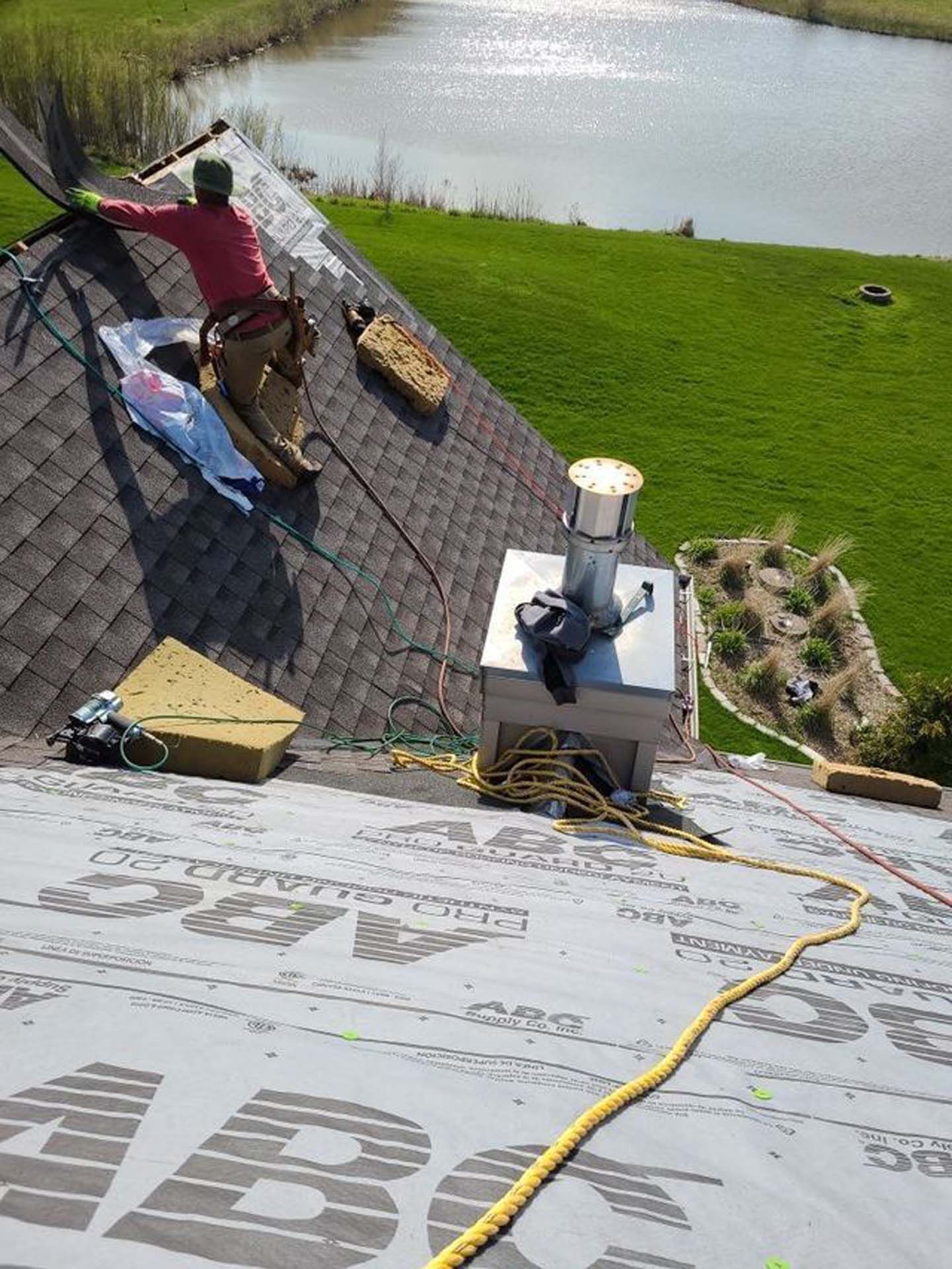 A worker installs shingles on a roof near a metal chimney vent, with a grassy area and pond in the background.