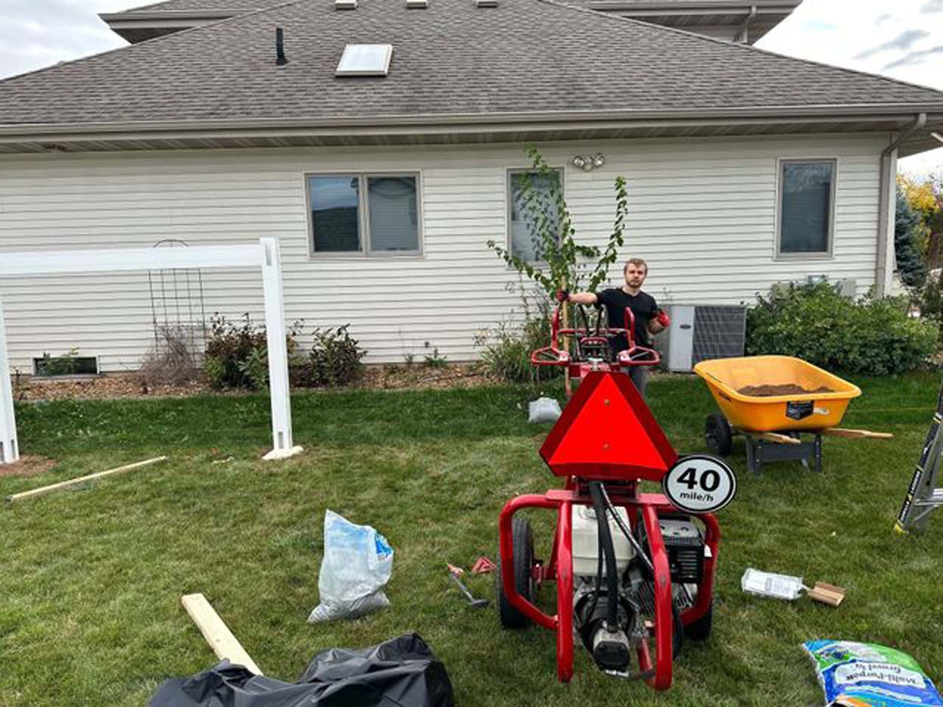 A man is standing in front of a house with a tractor and a wheelbarrow.