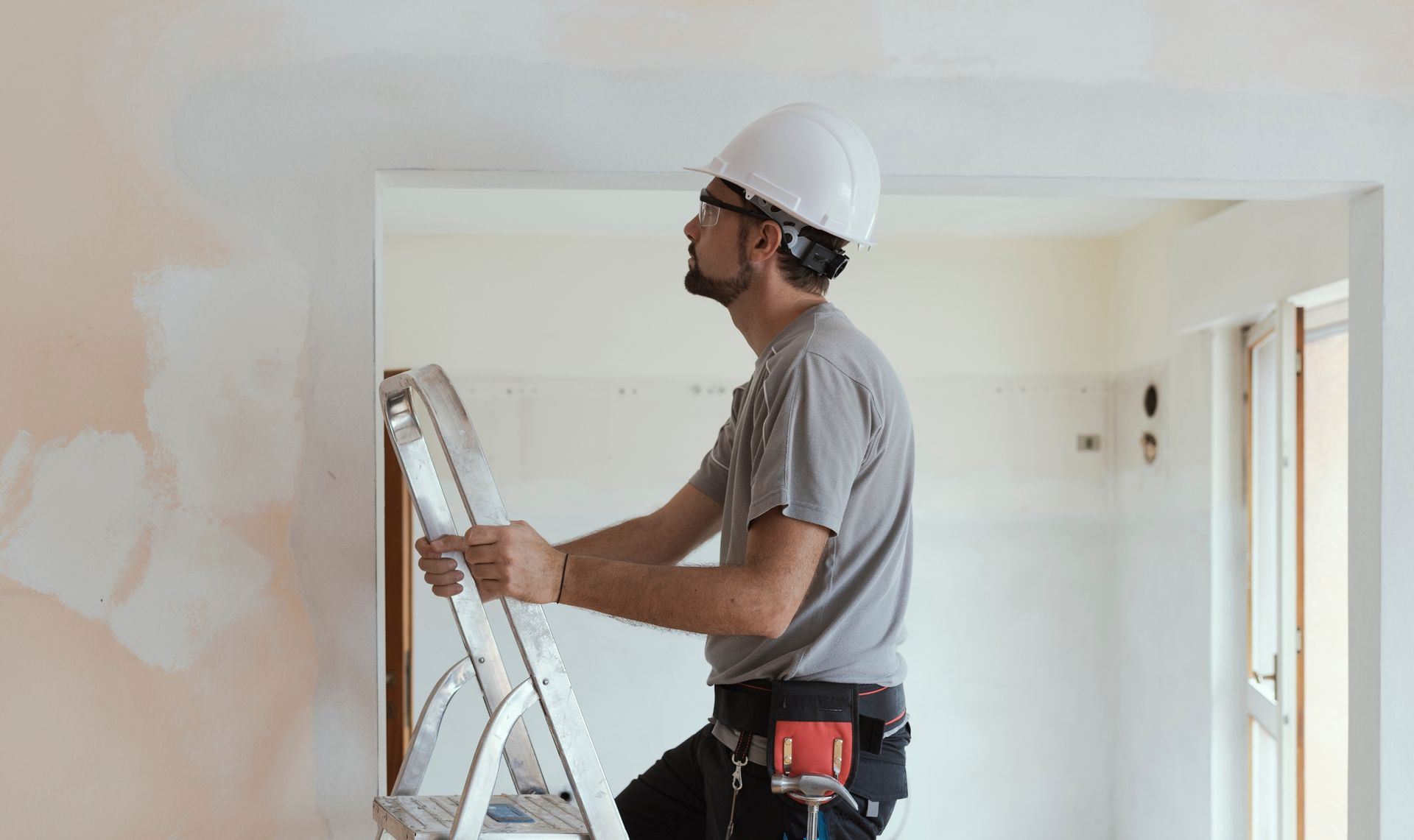 A person wearing a hard hat and tool belt stands on a step ladder, inspecting the frame of a doorway in a room.