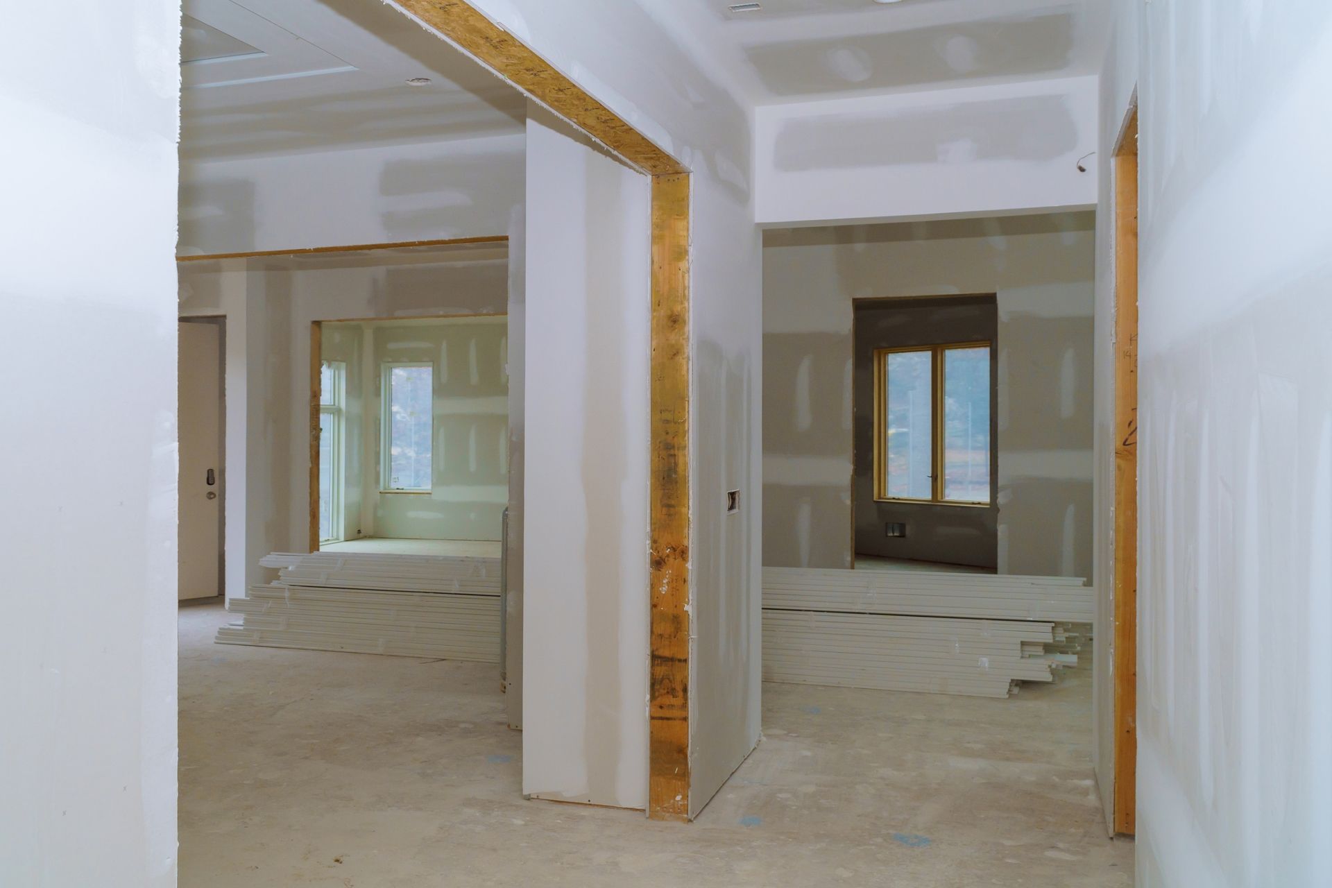 An interior view of a room under construction, showing bare drywall, wooden door frames, and window openings.