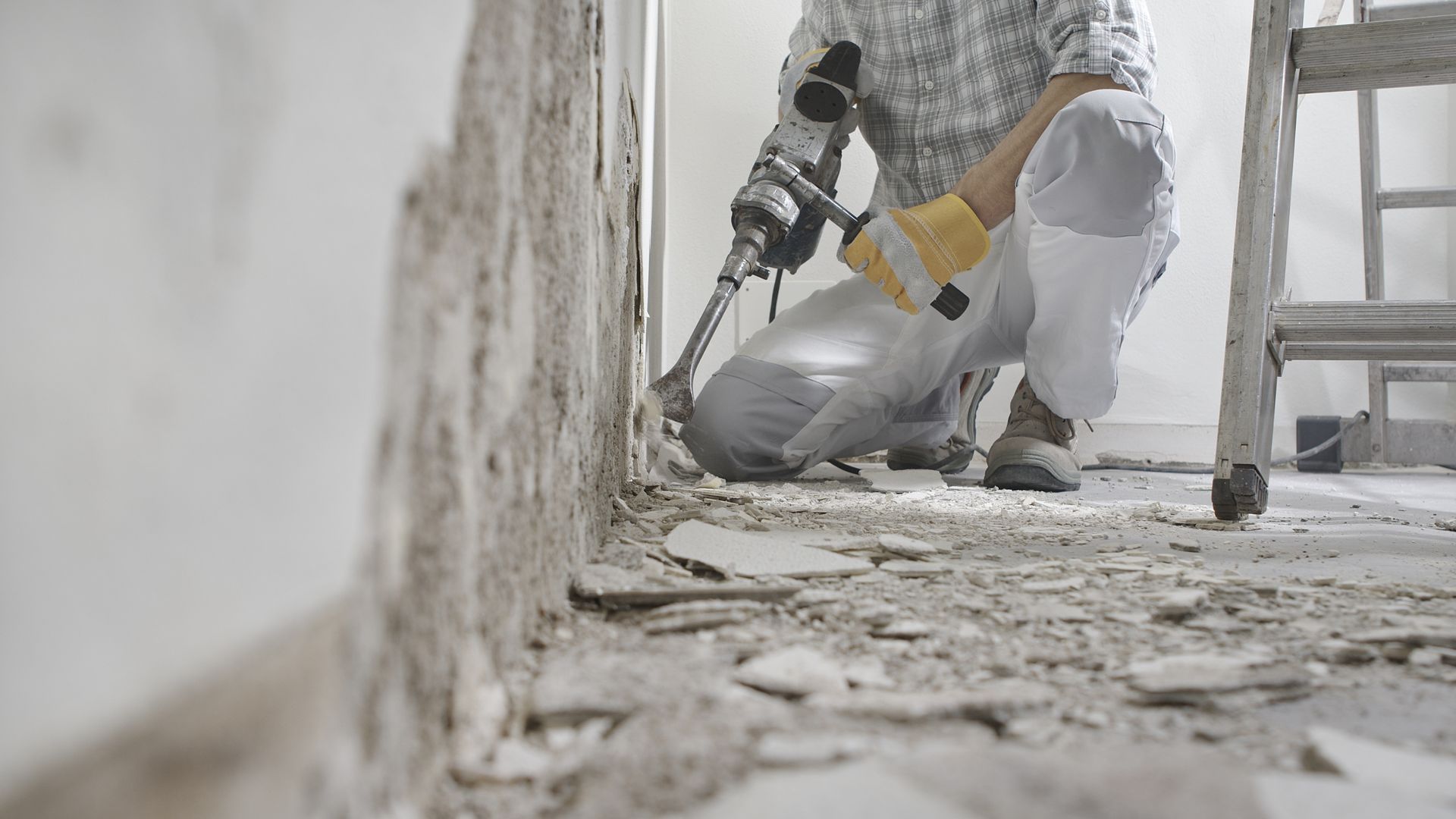 A worker in work clothes uses a power tool to remove plaster from a wall in a room under renovation.