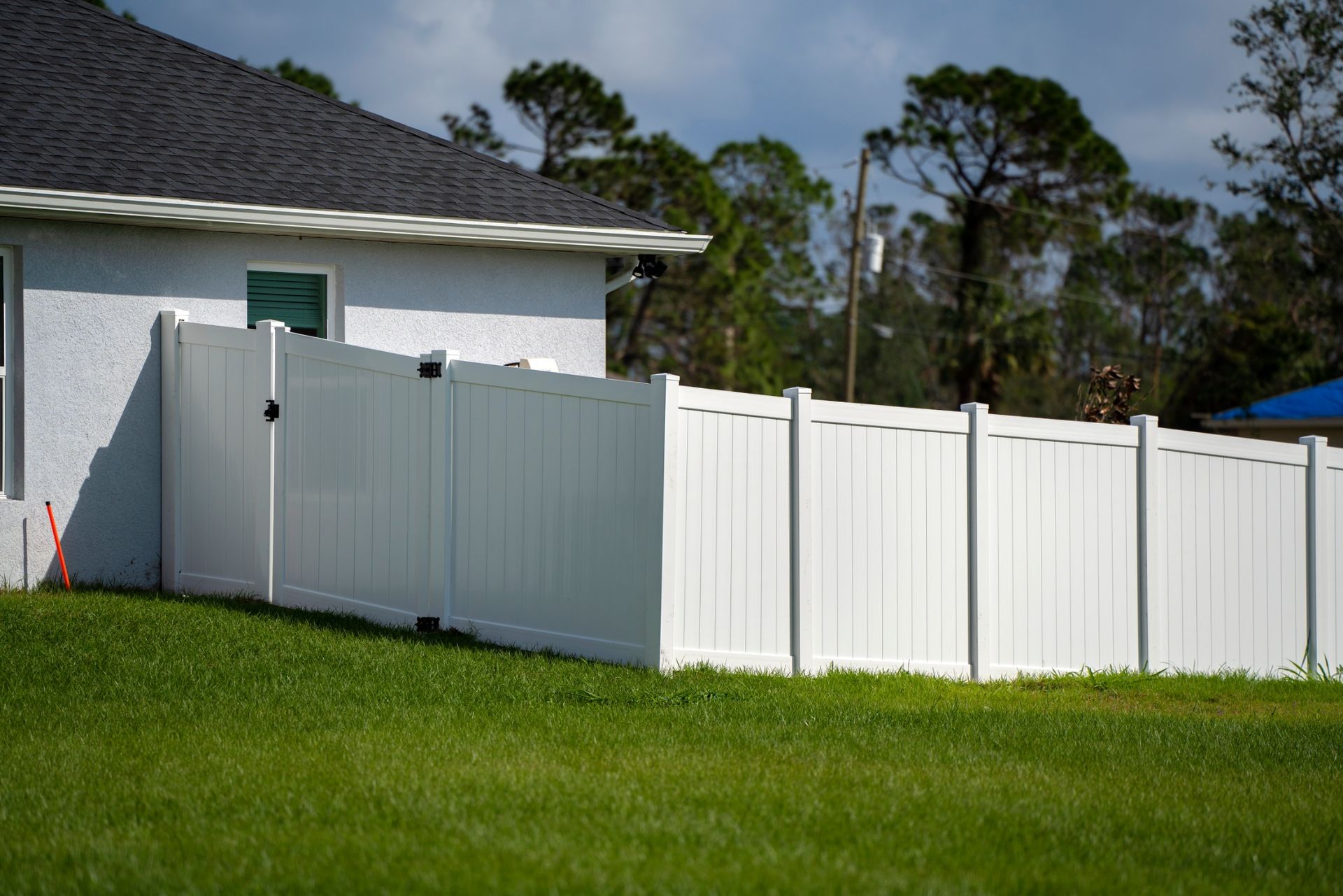 A white vinyl privacy fence with a closed gate runs along the side of a house on a green lawn against a sunny sky.
