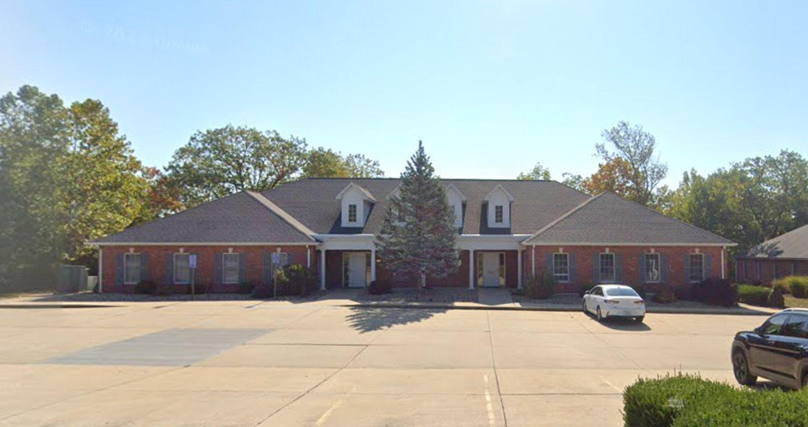 Two-story brick building with green roof, white columns, blue shutters, and address