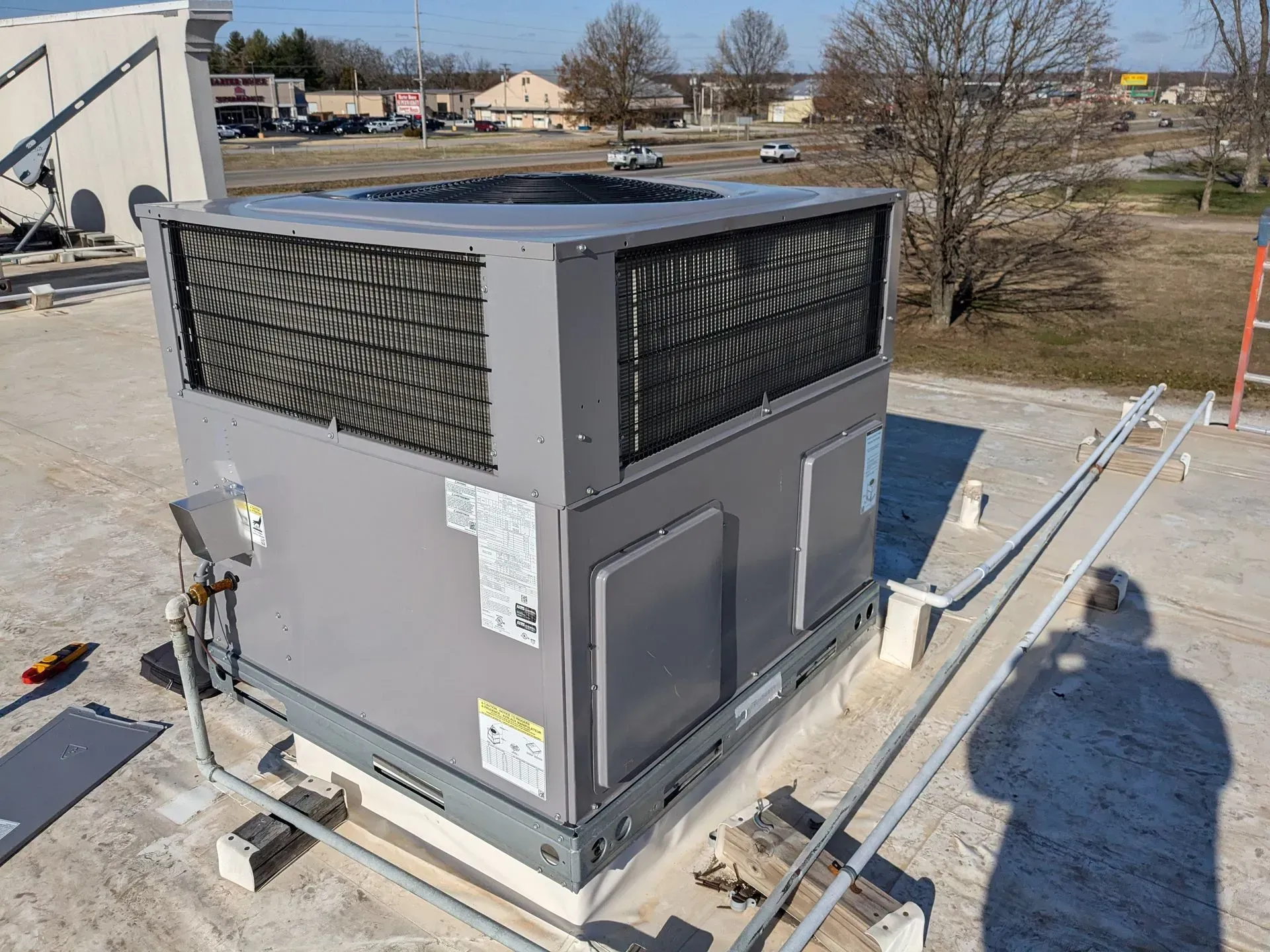 Rooftop HVAC unit, gray with black screens, sitting on a white building roof, with piping and ladder nearby.