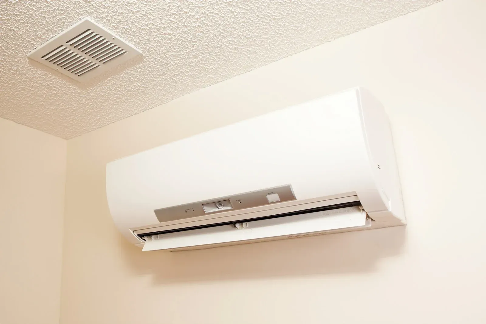 White air conditioning unit mounted on a cream-colored wall, with a ventilation grate in the ceiling above.