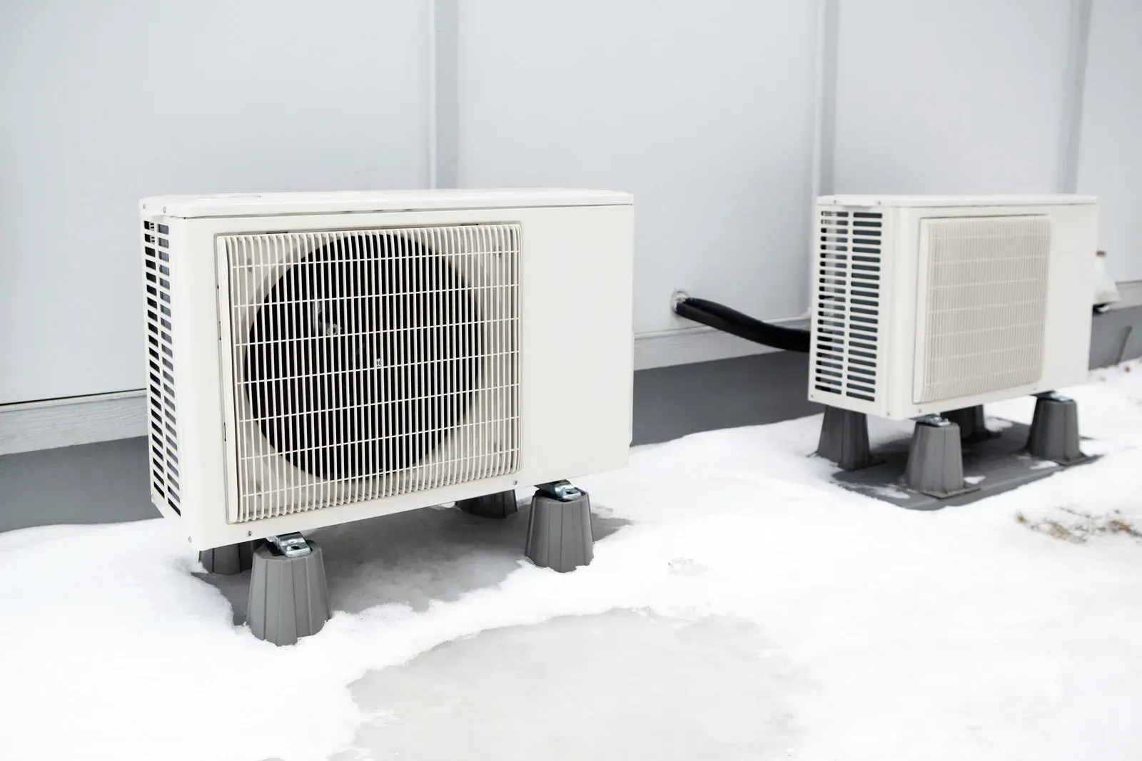 Two white air conditioning units on a snowy rooftop, elevated on black platforms.