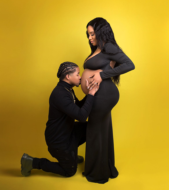 A couple in black outfits stands against a yellow backdrop, with one person kneeling and kissing the other's belly.