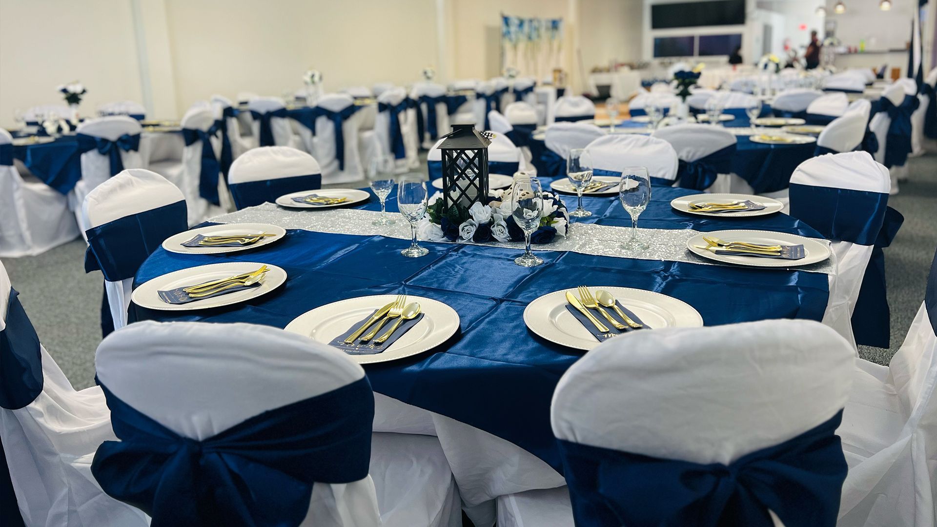 Tables set for a formal event with white tablecloths, navy blue runners, and chairs featuring white covers and blue sashes.