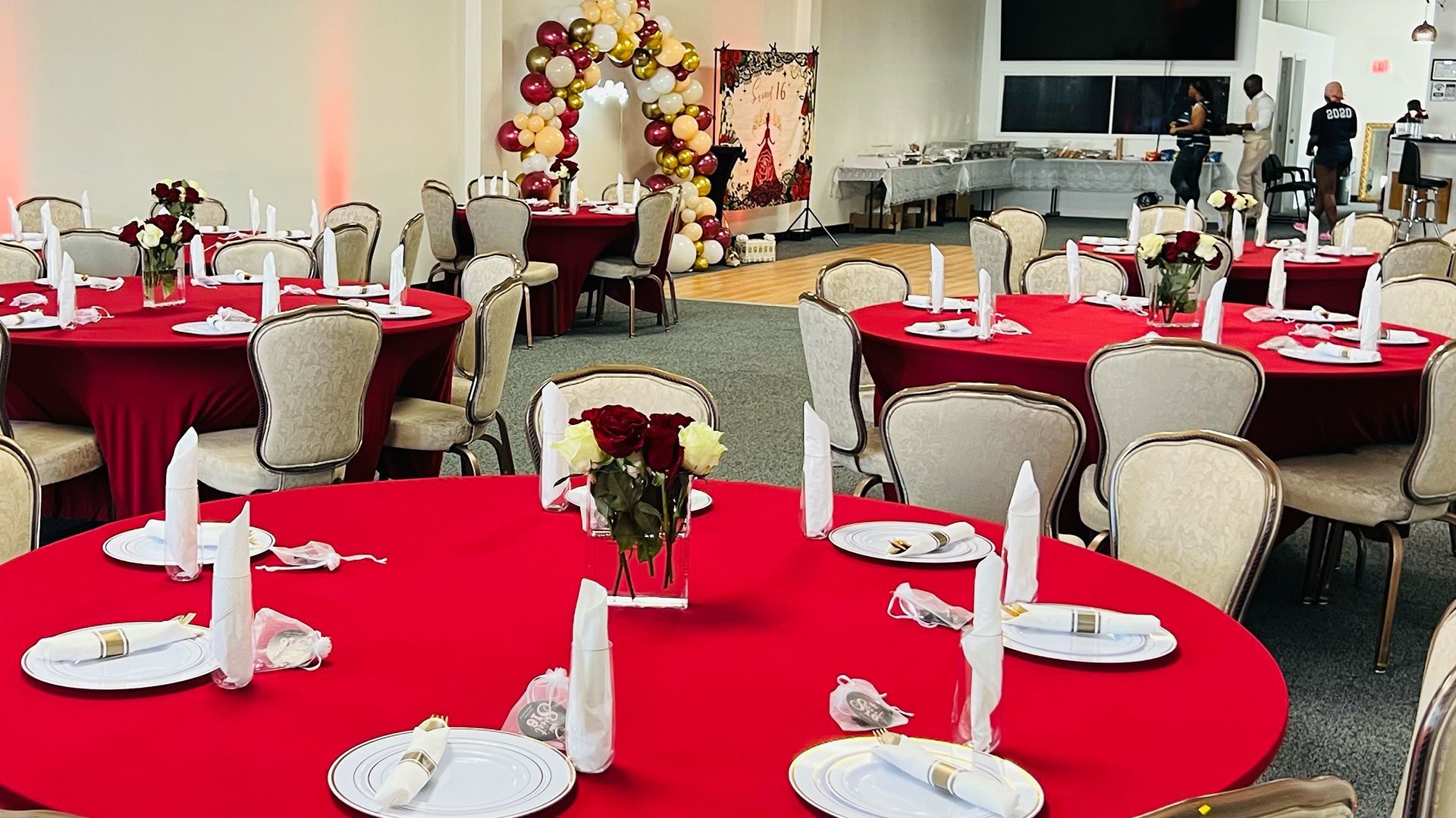 An event hall with round tables set with red tablecloths, white napkins, and centerpieces for a formal gathering.