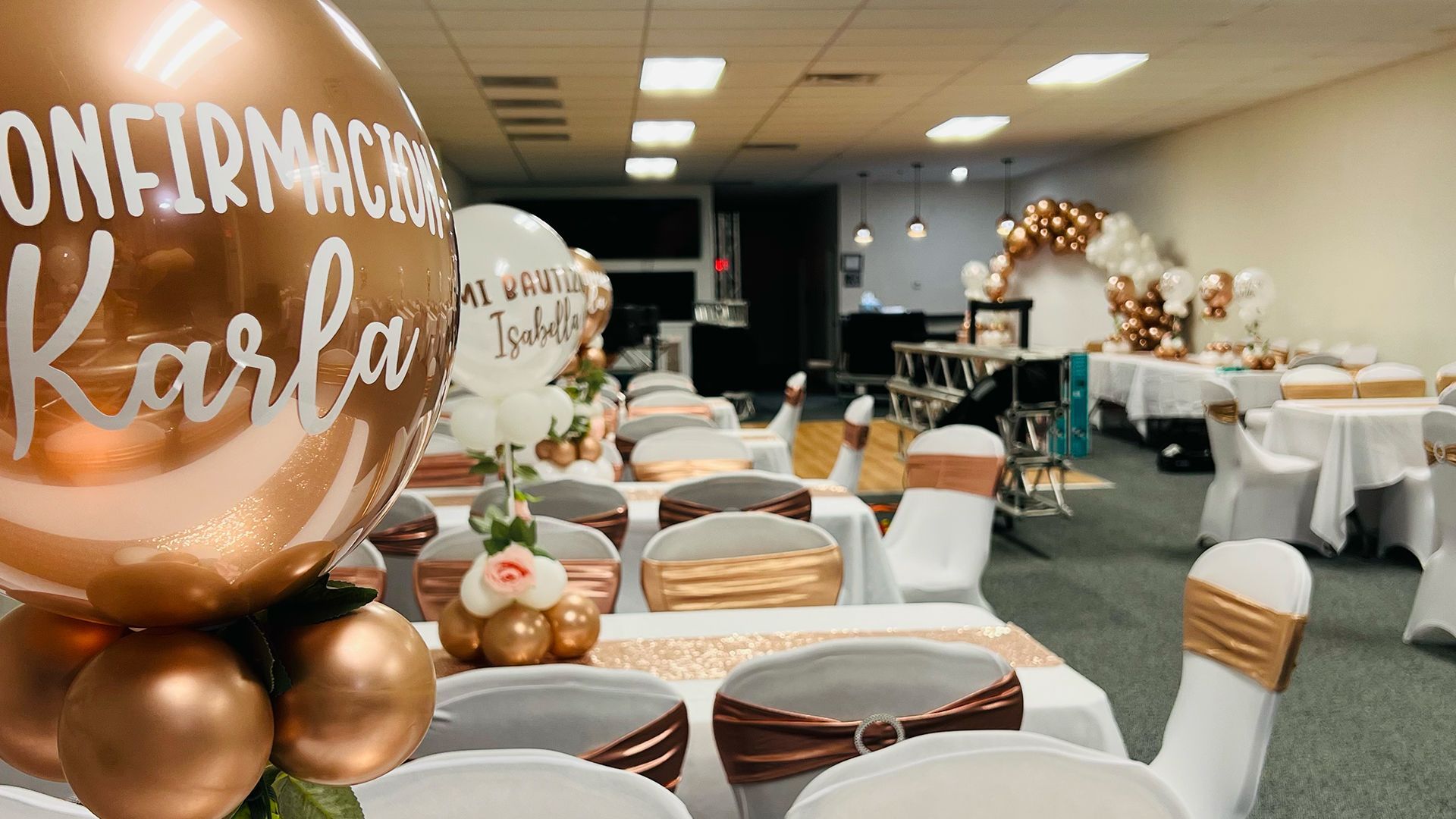 A decorated banquet hall with rose gold balloons and white chairs with matching sashes for a confirmation celebration.