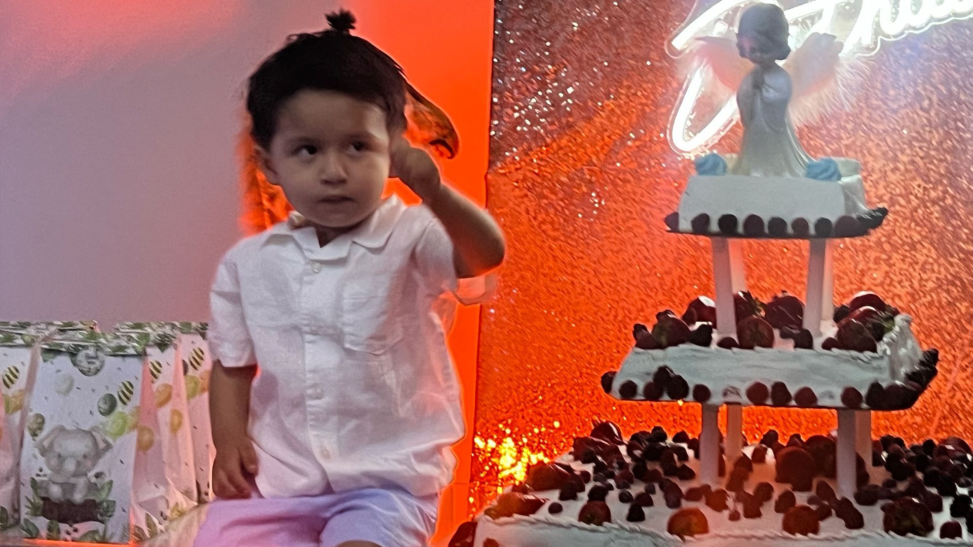 A person in a white shirt sits next to a tiered cake topped with a figurine against a bright orange, glittery backdrop.