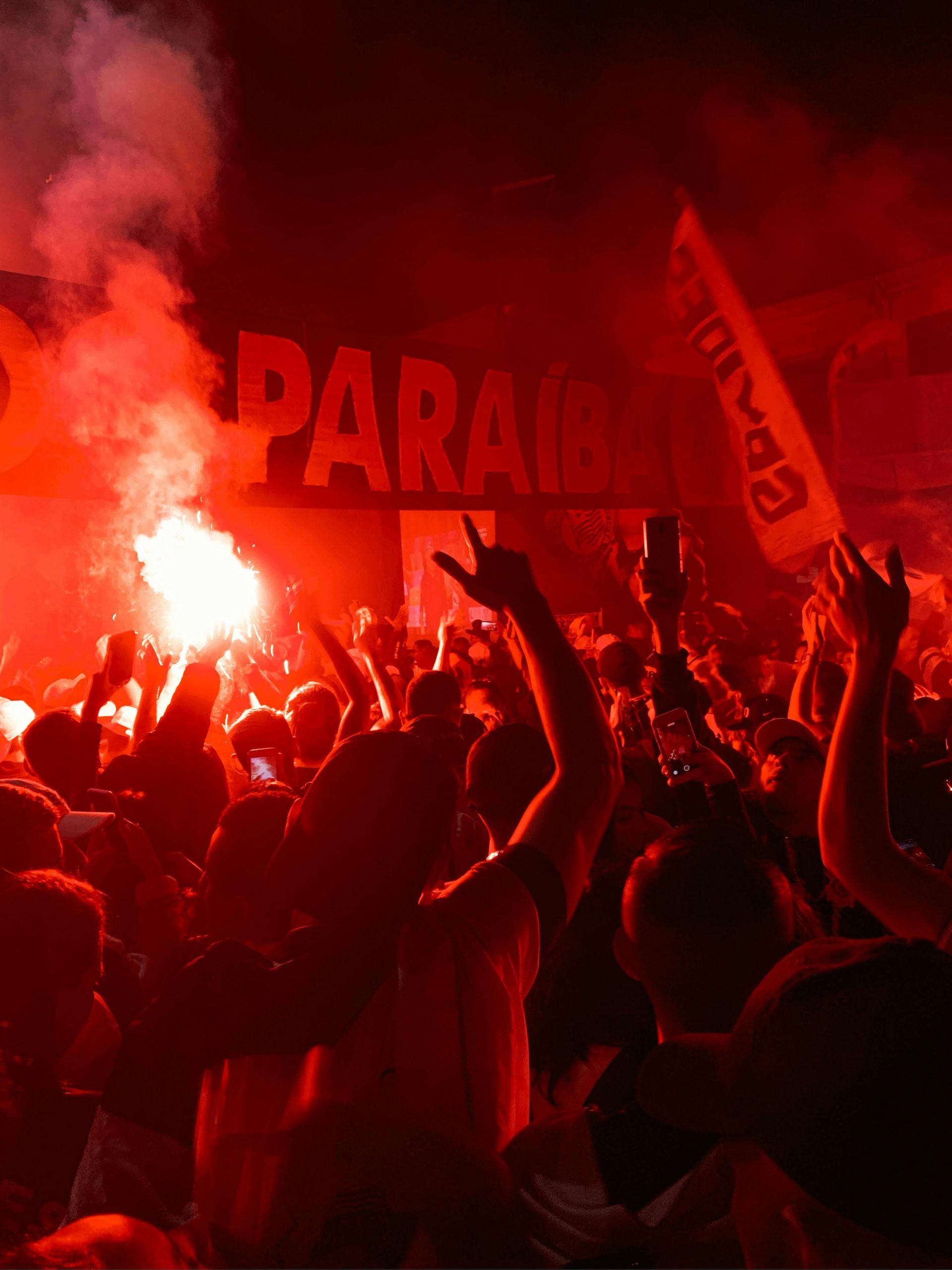 A crowd of people in a dark, red-lit space, with some holding hands up and a bright flare burning on the left.