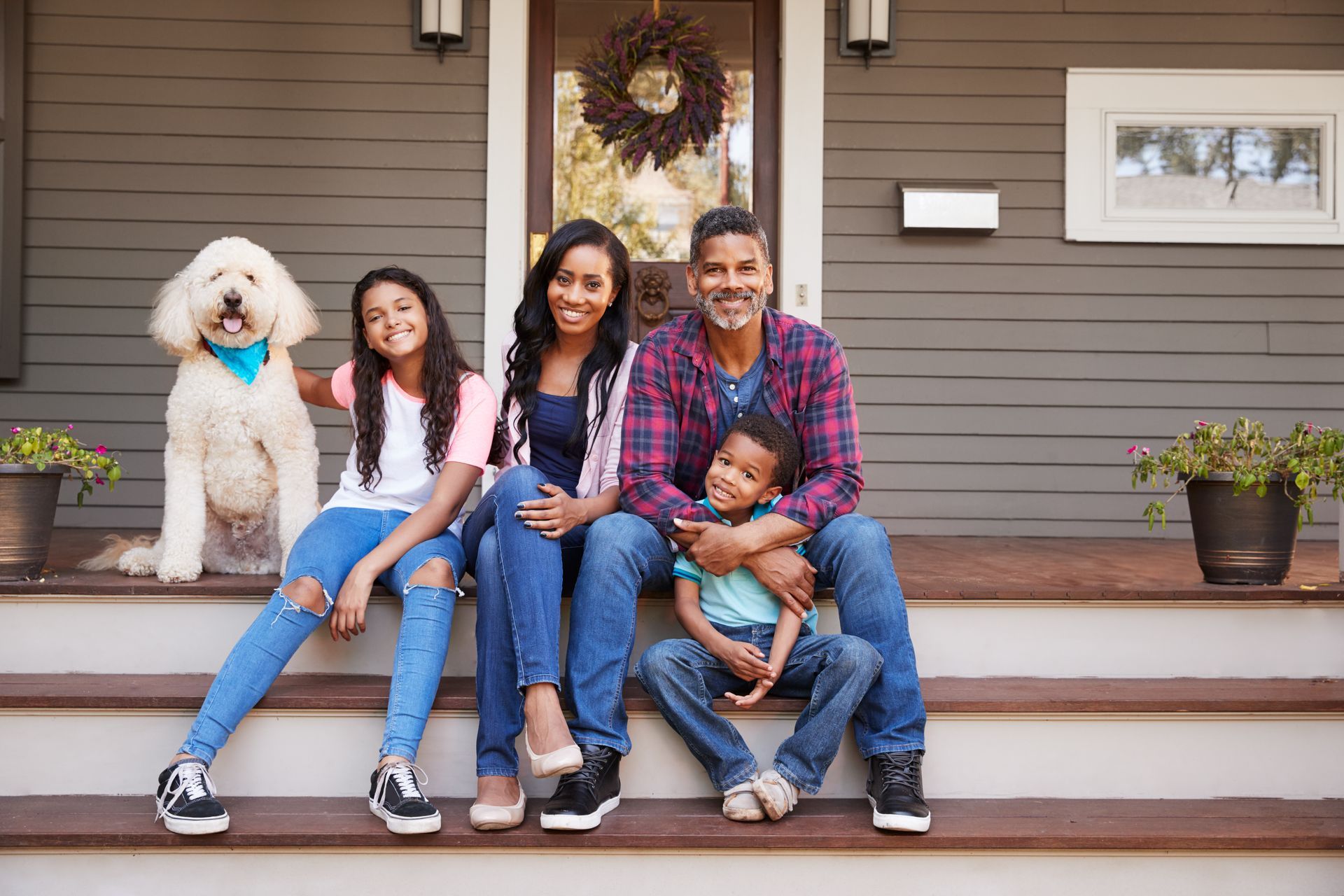 Family and fluffy dog sitting on front steps of a house with wreath and potted plants